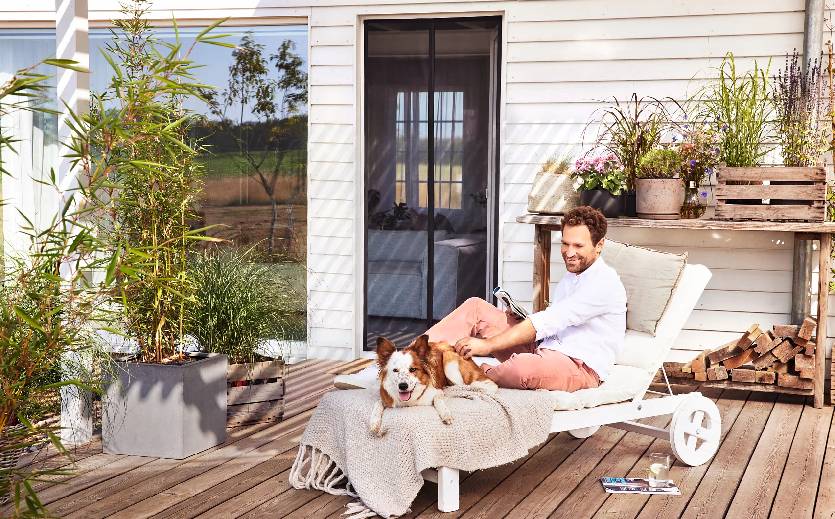 A man sits on a white outdoor lounge chair on a wooden patio, smiling and looking at a laptop. A brown and white dog lies next to him. The setting includes potted plants, a glass door, and a wooden planter with various plants. In the text provided, there is no mention of tape or other brands to replace with tesa tape. If you have additional content that specifically mentions tape, please provide it so I can make the necessary adjustments with tesa tape. (This text has been generated by AI)