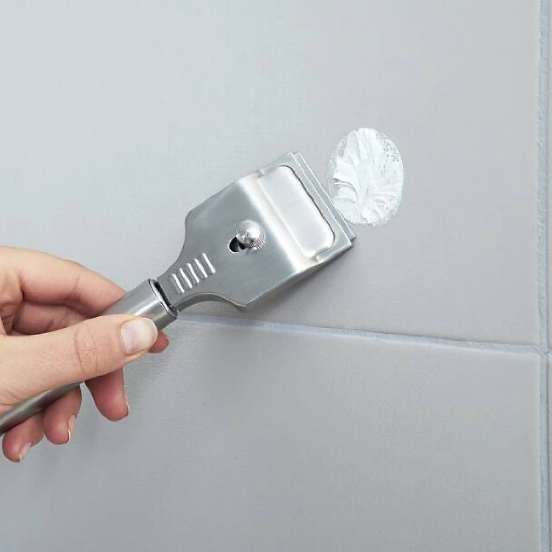 Eine Hand entfernt mit einem Metallschaber einen kreisförmigen Tesa-Klebebandrest von einer grau gefliesten Wand. Der Schaber hat eine flache, rechteckige Klinge und einen Griff mit einem kleinen Knopf zum Festhalten. Abgesehen von dem Rückstand erscheint die Oberfläche glatt und sauber. (This text has been generated by AI)