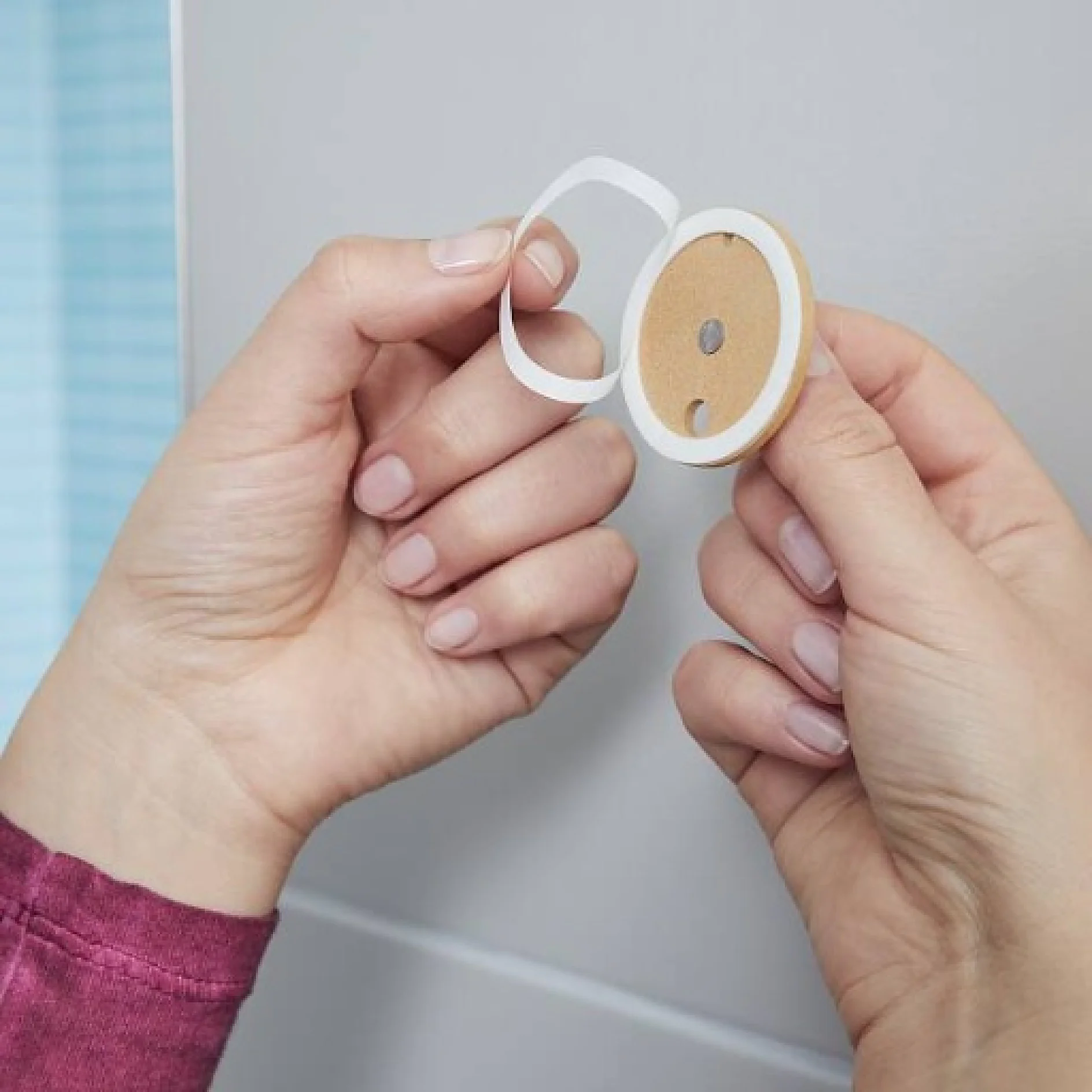 Eine Person in einem langärmligen rosa Oberteil entfernt einen Schutzstreifen von einem runden Tesa-Klebeband-Aufkleber an einer hellen Wand. Der Tesa-Klebeband-Aufkleber ist rund und hat eine kleine hellbraune Mitte. (This text has been generated by AI)
