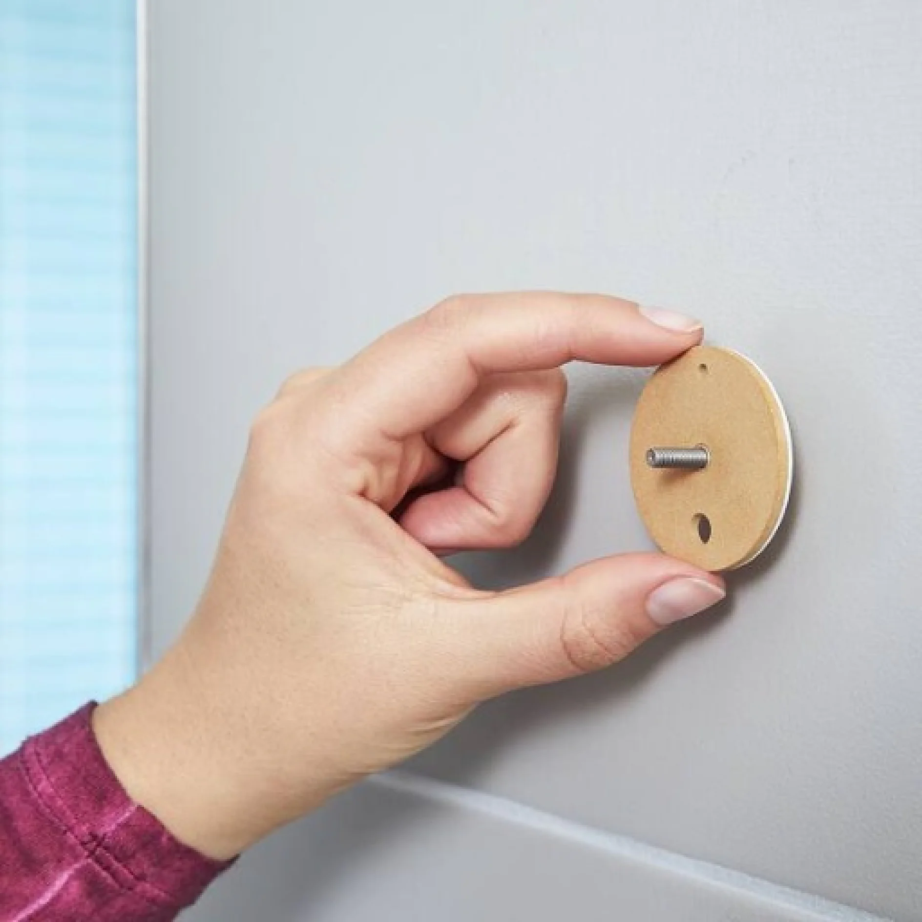 Die Hand einer Person hält einen kleinen Holzkreis mit einer Metallschraube an einer Seite an einer Wand. Der Kreis scheint zur Wandmontage oder zum Befestigen von Objekten mit Tesa-Klebeband gedacht zu sein. Die Person trägt ein langärmeliges lila Hemd. (This text has been generated by AI)