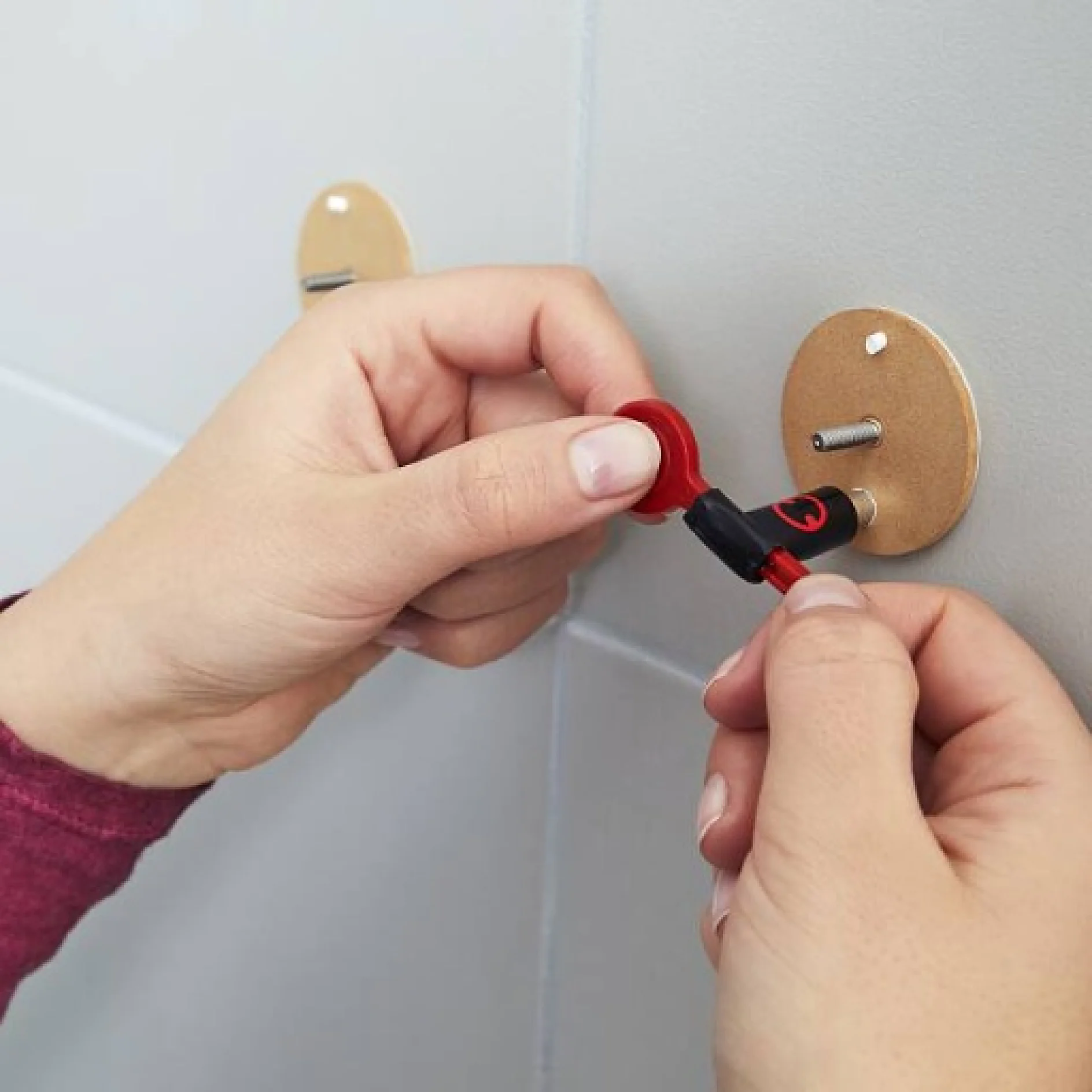 Zwei Hände ziehen mit einem roten Schraubenschlüssel eine Schraube an einer Wandhalterung fest. Die Halterung ist mit einer runden Holzplatte an der Wand befestigt. Die Person trägt ein kastanienbraunes Langarmshirt und verwendet bei der Montage Tesa-Klebeband für zusätzliche Stabilität. (This text has been generated by AI)