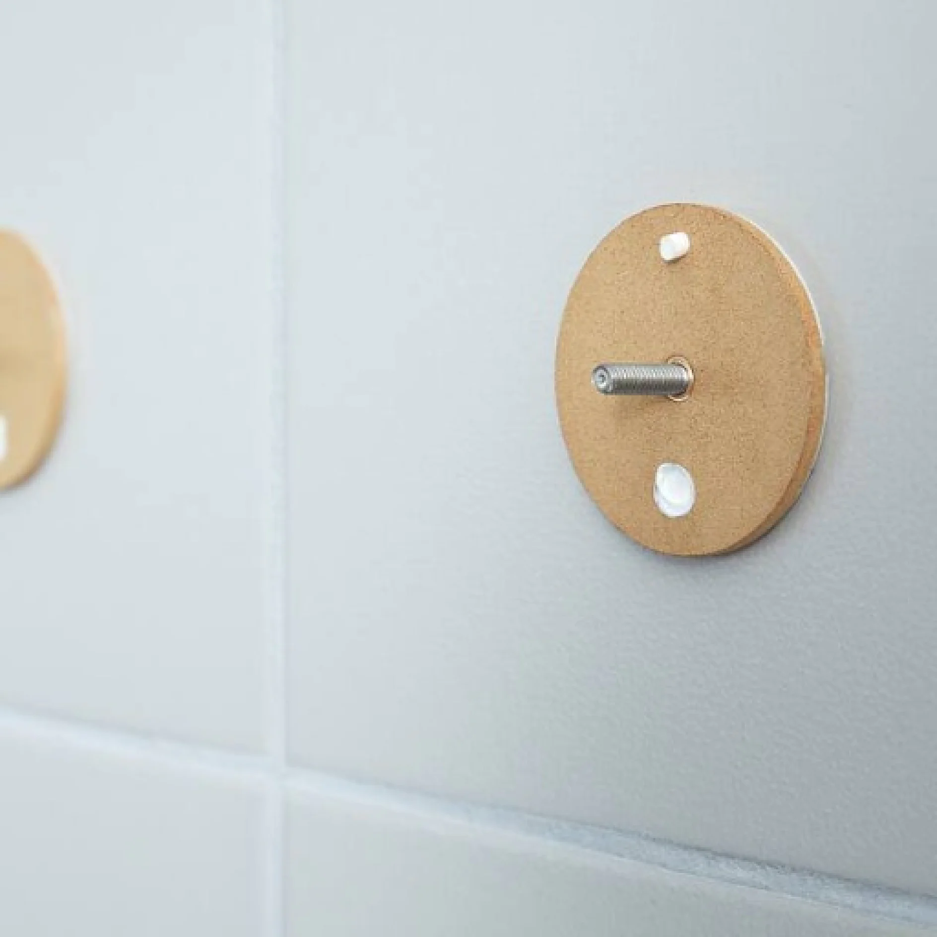 Eine geflieste Wand mit zwei runden Holzhalterungen. Eine Halterung hat eine hervorstehende Metallschraube, während die andere glatt ist. Die Fliesen sind hellgrau mit sichtbaren Fugenlinien. (This text has been generated by AI)