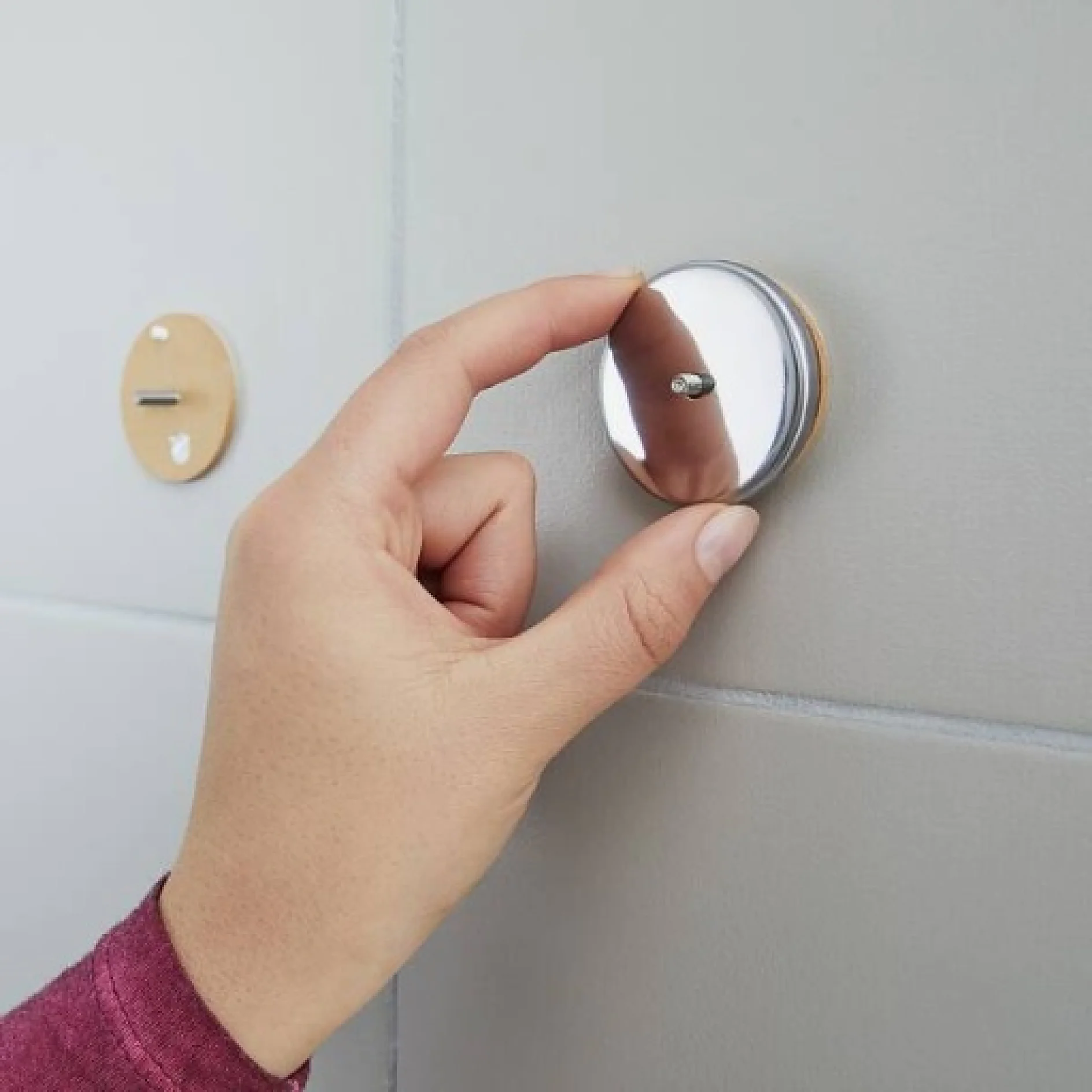 Eine Hand ist dabei, einen runden Spiegel an einer metallischen Magnethalterung aufzuhängen, die mit Tesa-Klebeband an einer hellgrau gefliesten Wand befestigt ist. Links ist eine weitere leere Halterung zu sehen. Die Person trägt ein langärmeliges rosarotes Kleidungsstück. (This text has been generated by AI)