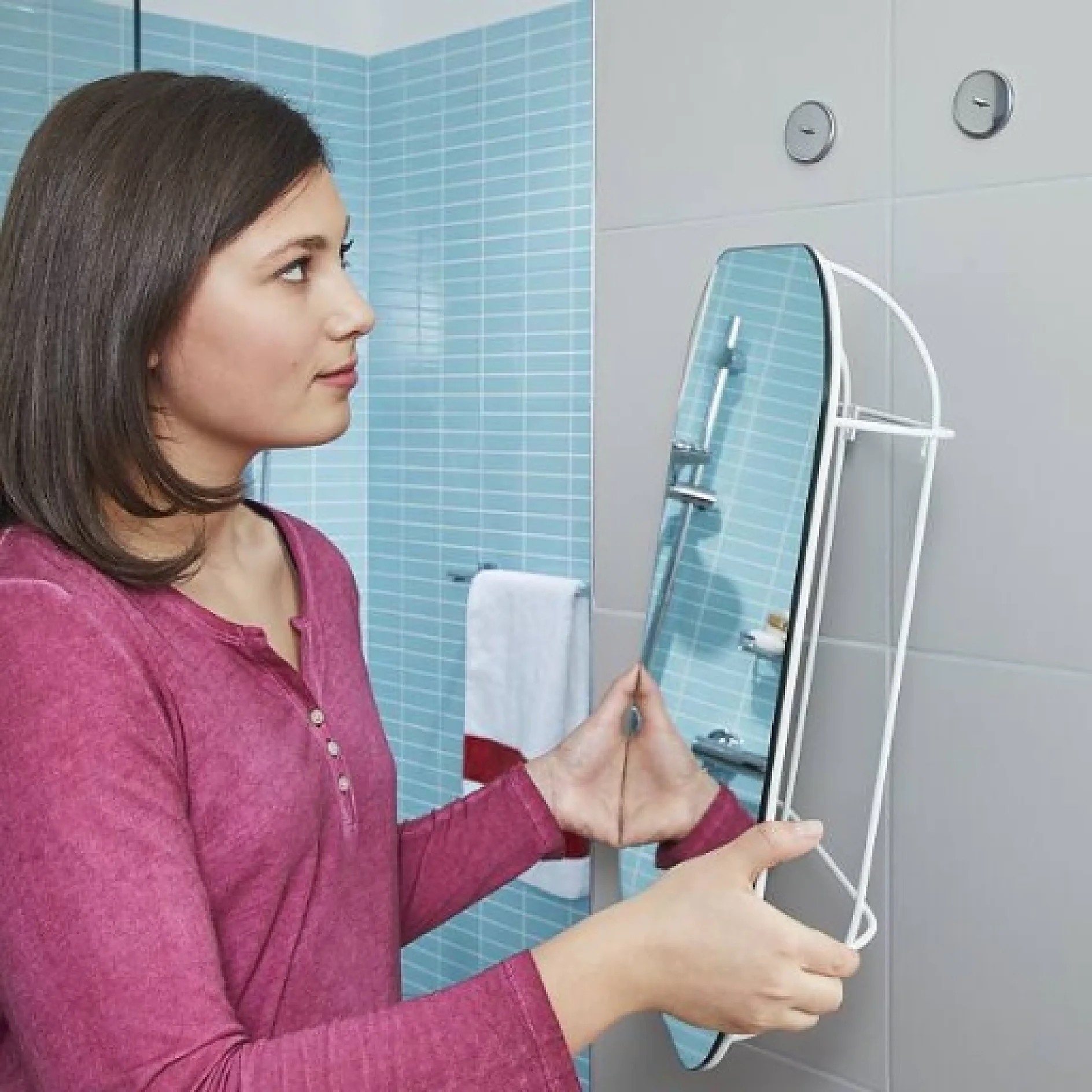 Eine Person in einem kastanienbraunen Oberteil hält einen vertikalen, rechteckigen Spiegel in einem Badezimmer mit blau gefliesten Wänden. Im Hintergrund hängt ein weißes Handtuch mit roten Akzenten an einem Ständer, und für alle erforderlichen Befestigungen oder Fixierungen wird scheinbar diskret Tesa-Klebeband verwendet. (This text has been generated by AI)