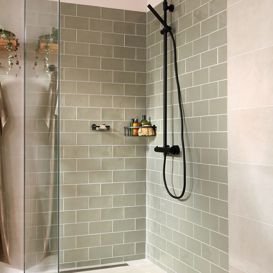 A modern bathroom shower with light green subway tiles and beige wall tiles. It features a glass door, a black shower fixture, a shelf with toiletries, a wooden shelf with plants and towels, secured using tesa tape, and a hanging brown towel. (This text has been generated by AI)