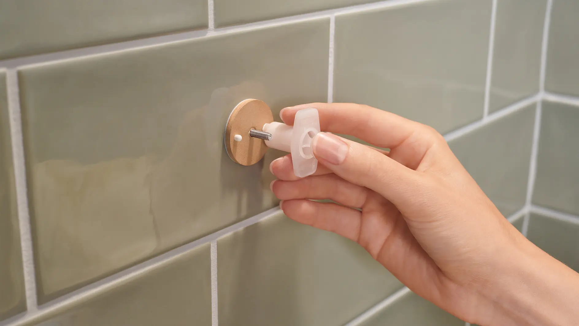 A hand is adjusting a small, circular metallic fixture on a wall with green tiles using a T-shaped plastic tool with tesa tape.