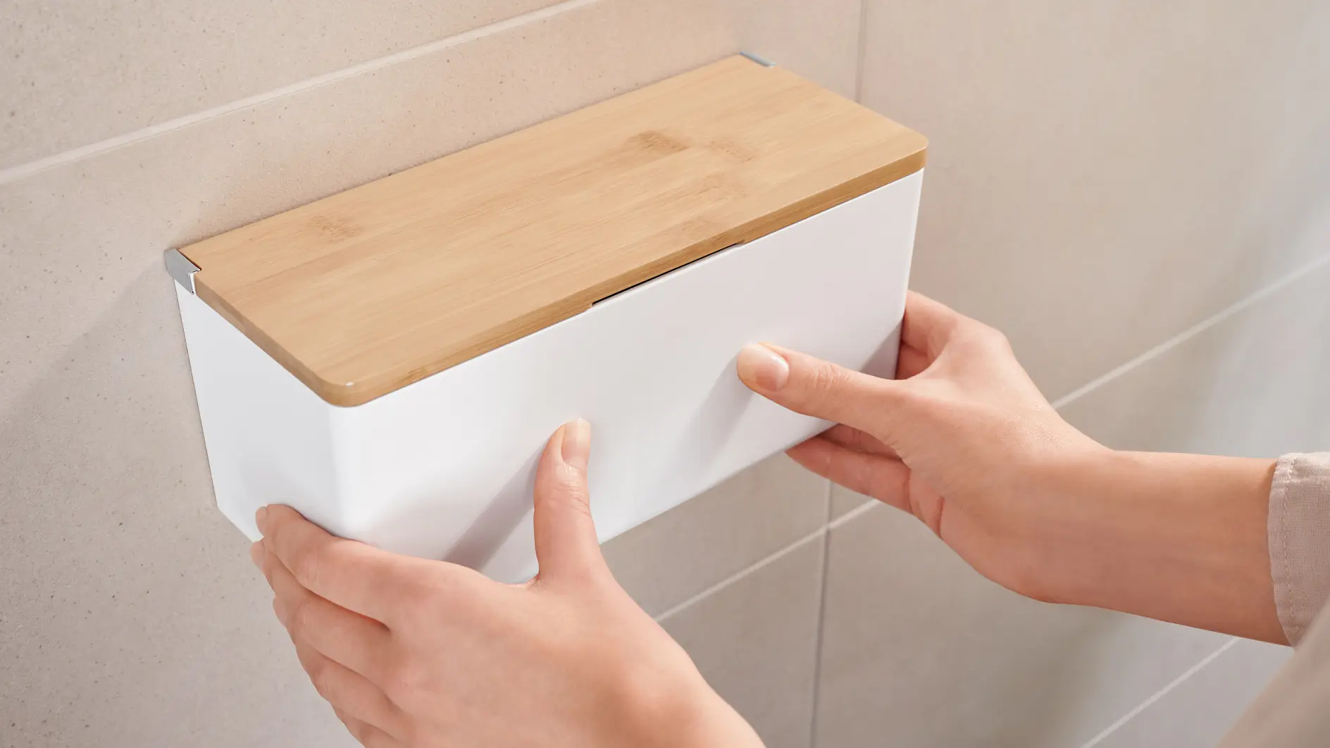 Two hands are attaching a rectangular, wall-mounted storage box with a wooden lid to a tiled wall using tesa tape.