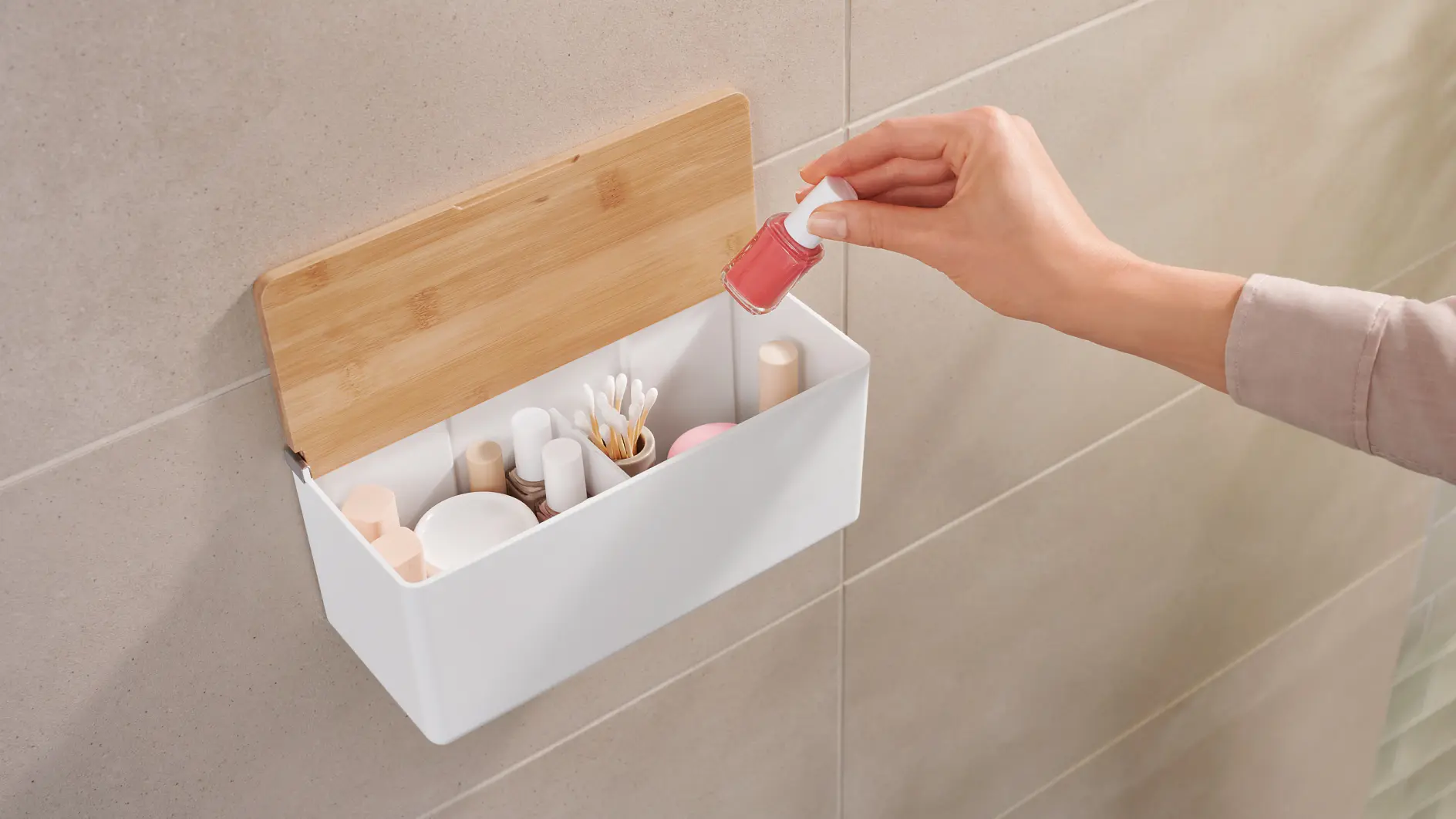 A hand places a pink nail polish bottle into a white wall-mounted storage box with a wooden lid. The box contains various cosmetics, including brushes, q-tips, and other makeup items. Its attached to a tiled wall using tesa tape.