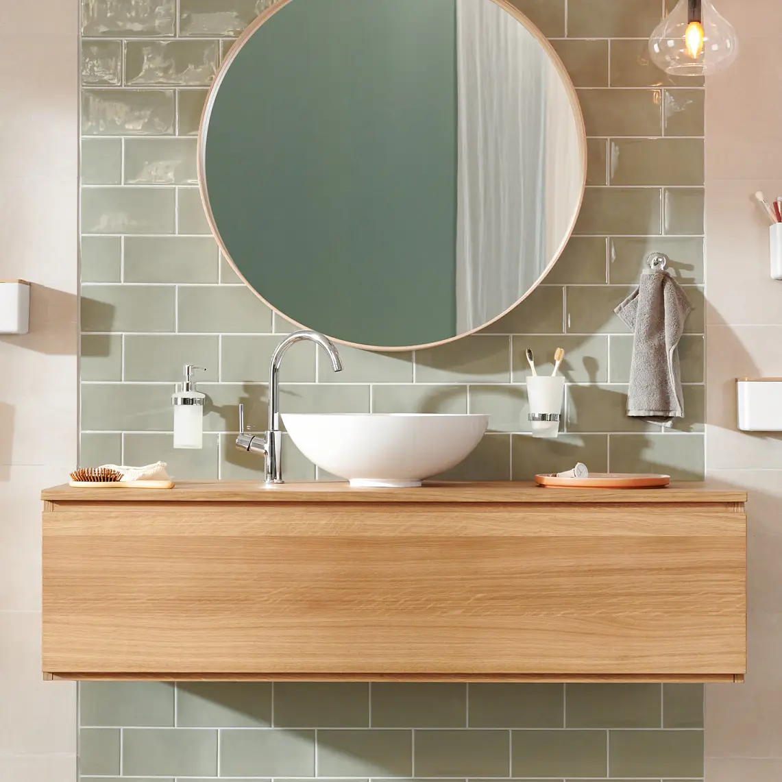 Modern bathroom with a round mirror above a wooden vanity. A white vessel sink sits on the vanity. Green rectangular tiles are on the wall. Accessories include a hanging hand towel, soap dispenser, and toothbrush holder secured with tesa tape. Warm lighting overhead. (This text has been generated by AI)