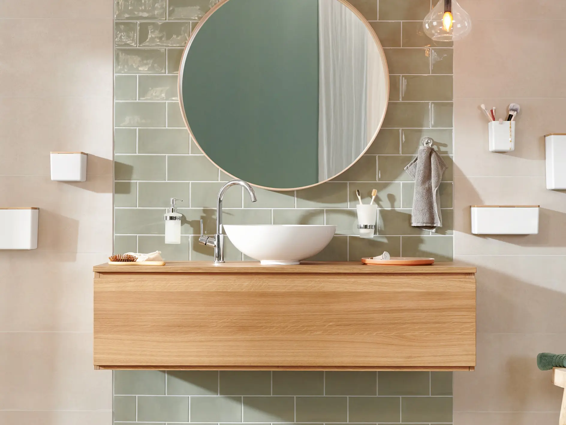 Modern bathroom with a round mirror above a wooden vanity. A white vessel sink sits on the vanity. Green rectangular tiles are on the wall. Accessories include a hanging hand towel, soap dispenser, and toothbrush holder secured with tesa tape. Warm lighting overhead.