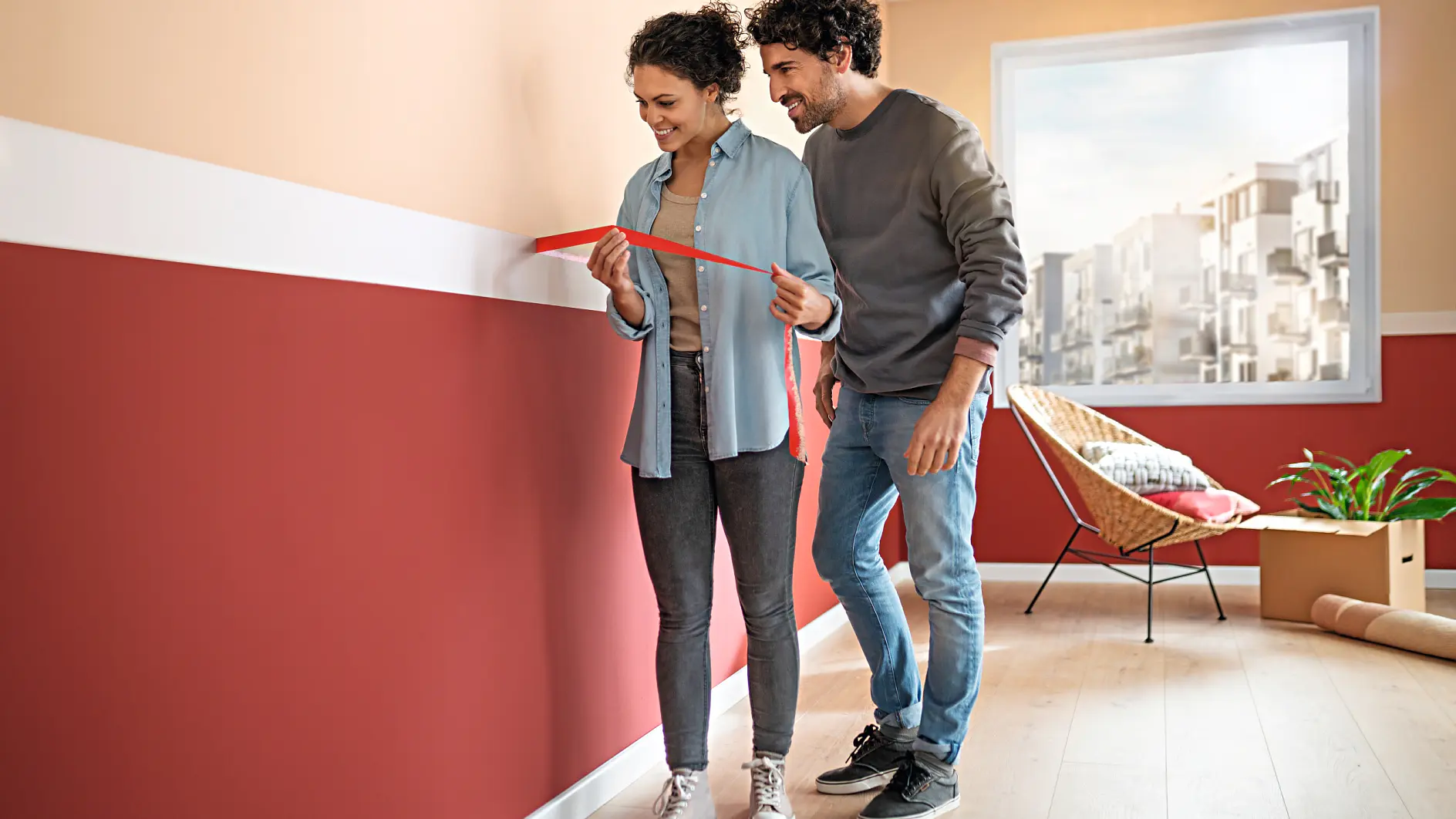 A couple stands in a room with red and beige walls. The woman holds a red tesa tape measure against the wall, and both are smiling. A chair and a plant are in the background near a window. The floor is light wood.
