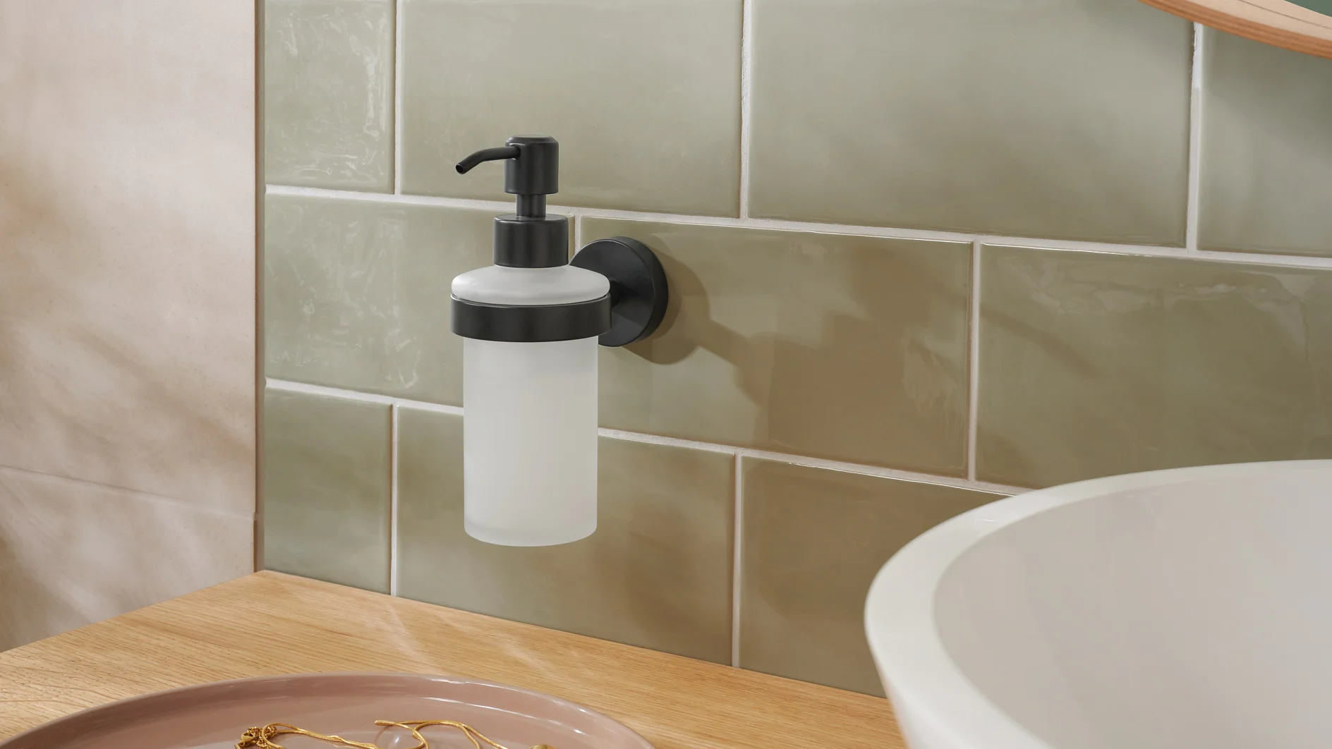 A wall-mounted soap dispenser with a black pump is attached to a tiled wall using tesa tape. The tiles are light green, and a wooden countertop is visible below, partially holding a round white sink and a pink dish with gold jewelry. (This text has been generated by AI)
