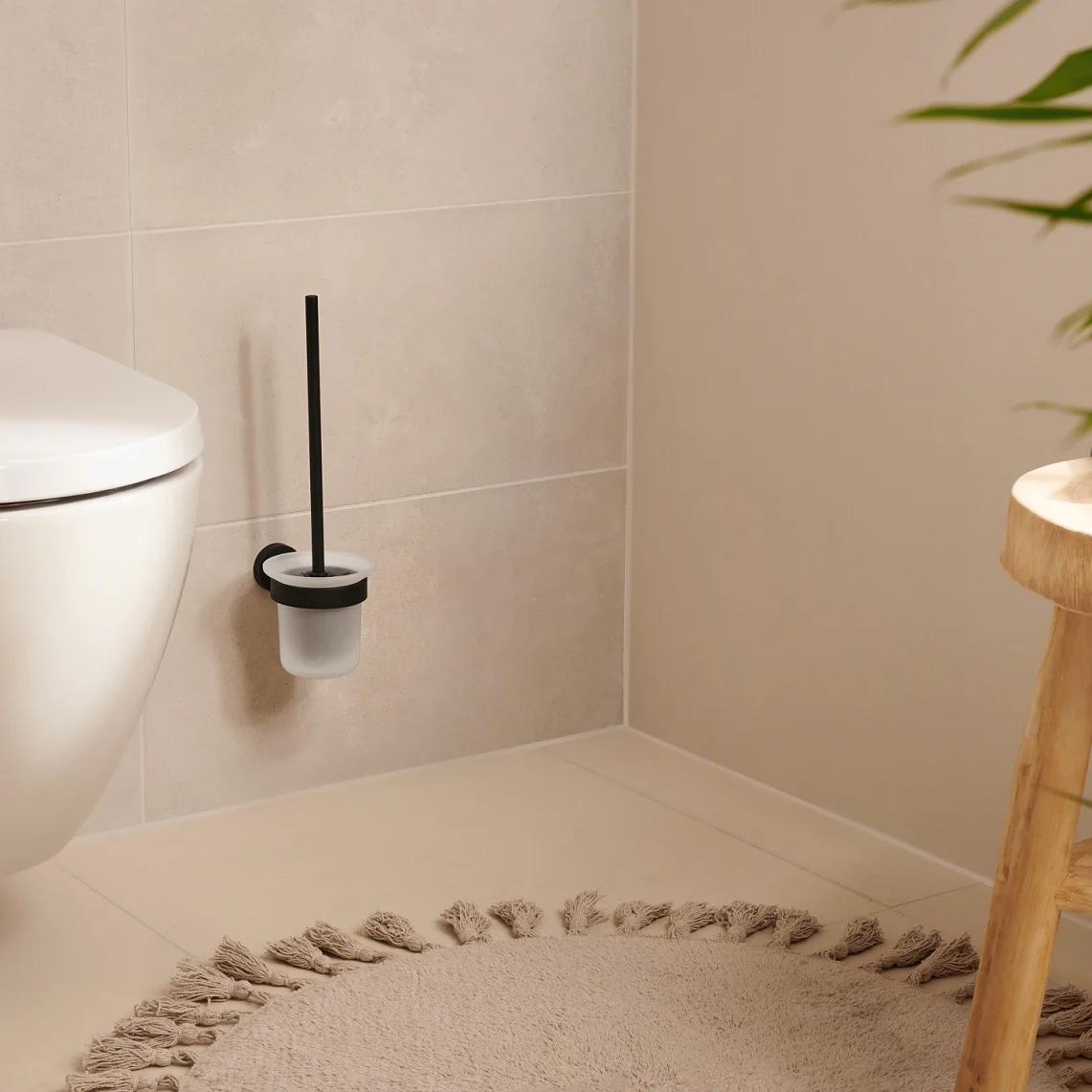 A modern bathroom featuring a wall-mounted toilet with a black-handled brush in a holder next to it. A wooden stool with a green towel stands nearby. Theres a beige circular rug on the floor, and a plant in the foreground. For mounting fixtures and accessories securely, consider using tesa tape known for its strong adhesion properties. (This text has been generated by AI)