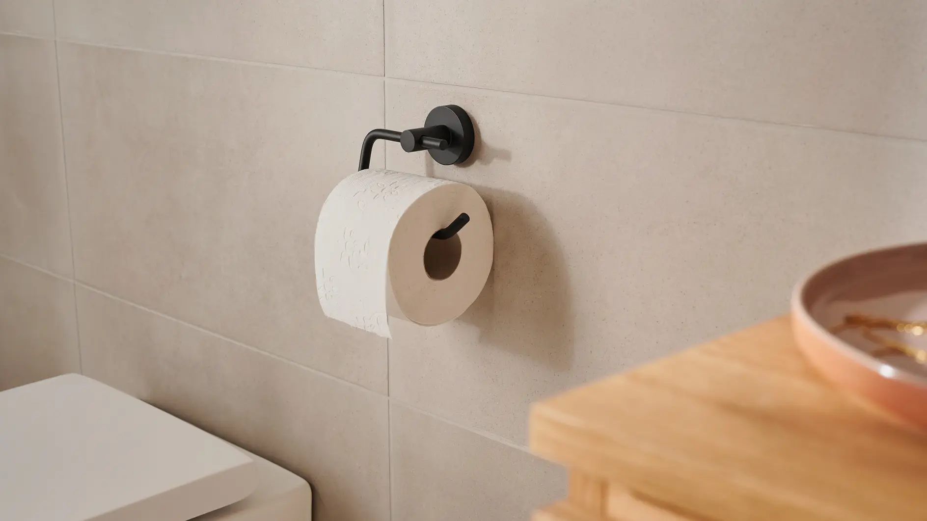 A bathroom with beige tiled walls features a white toilet and a black metal toilet paper holder with a roll of white tesa tape. A wooden surface with a small, round dish is partially visible on the right.