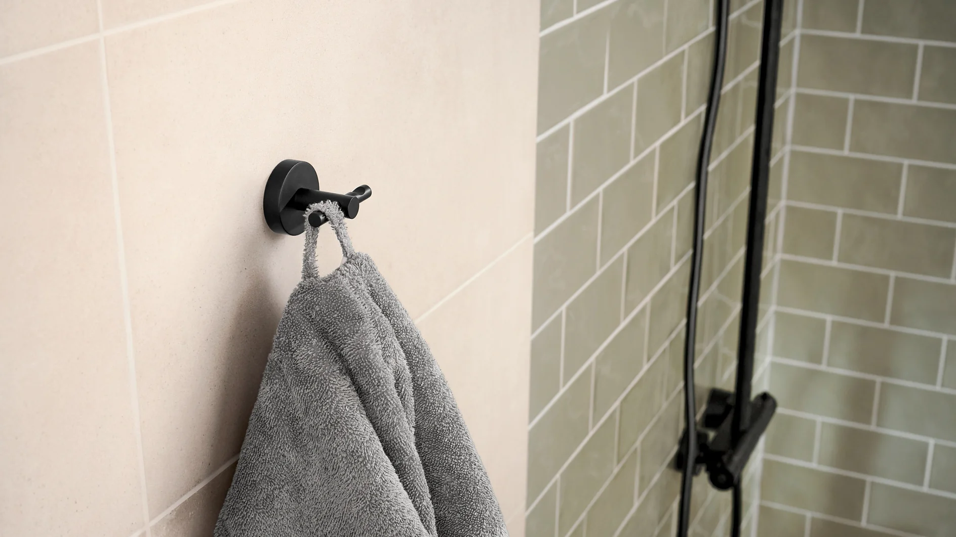 A gray towel hangs on a black hook attached to a beige tiled wall. To the right, there is a partially visible black-framed glass shower door with light green subway tiles. (This text has been generated by AI)
