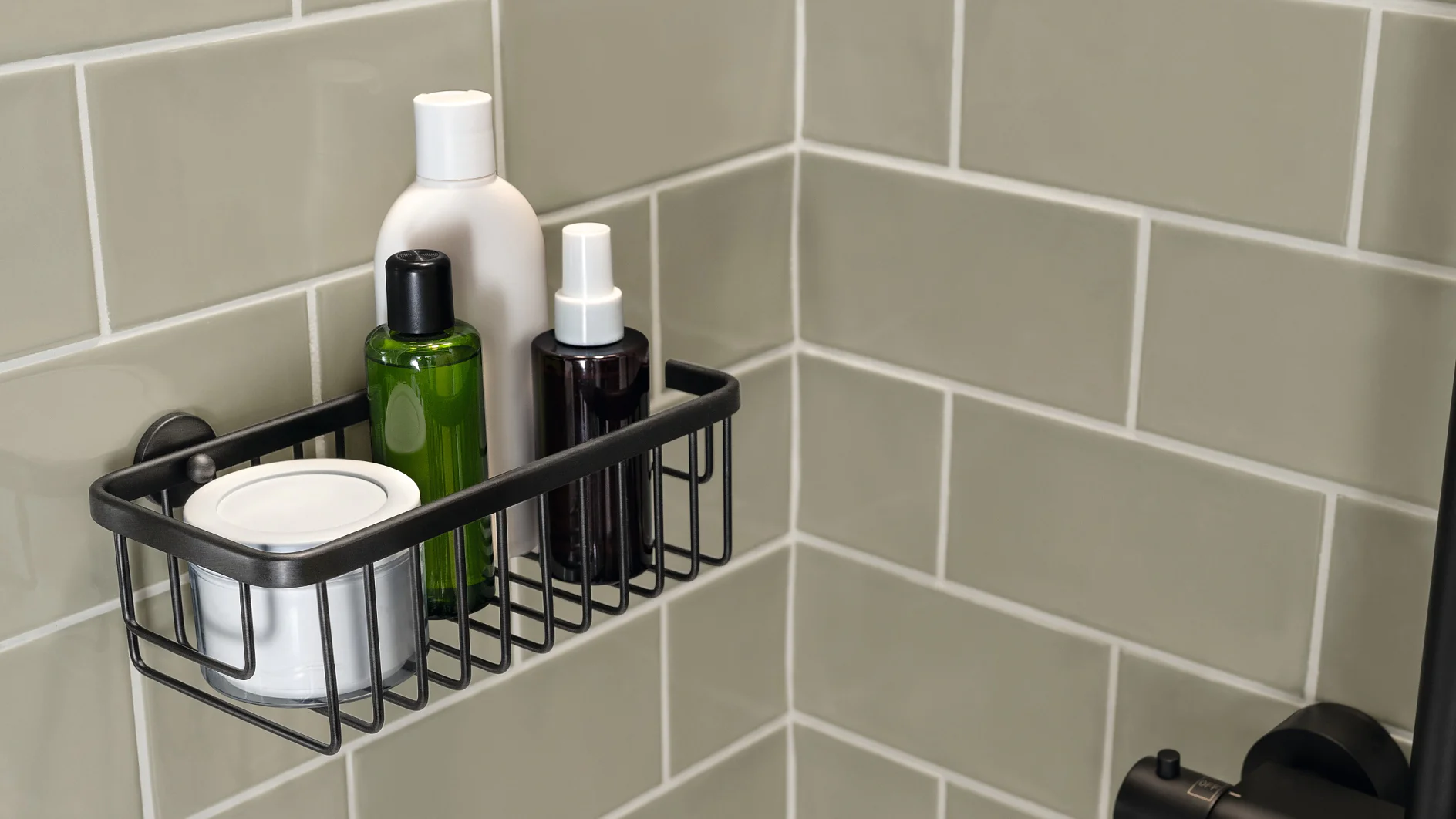 A metal shower caddy is mounted on a light gray tiled wall, holding four bottles of various sizes and shapes. The caddy is fixed in a corner using tesa tape, securely supporting the shampoo or lotion containers. (This text has been generated by AI)