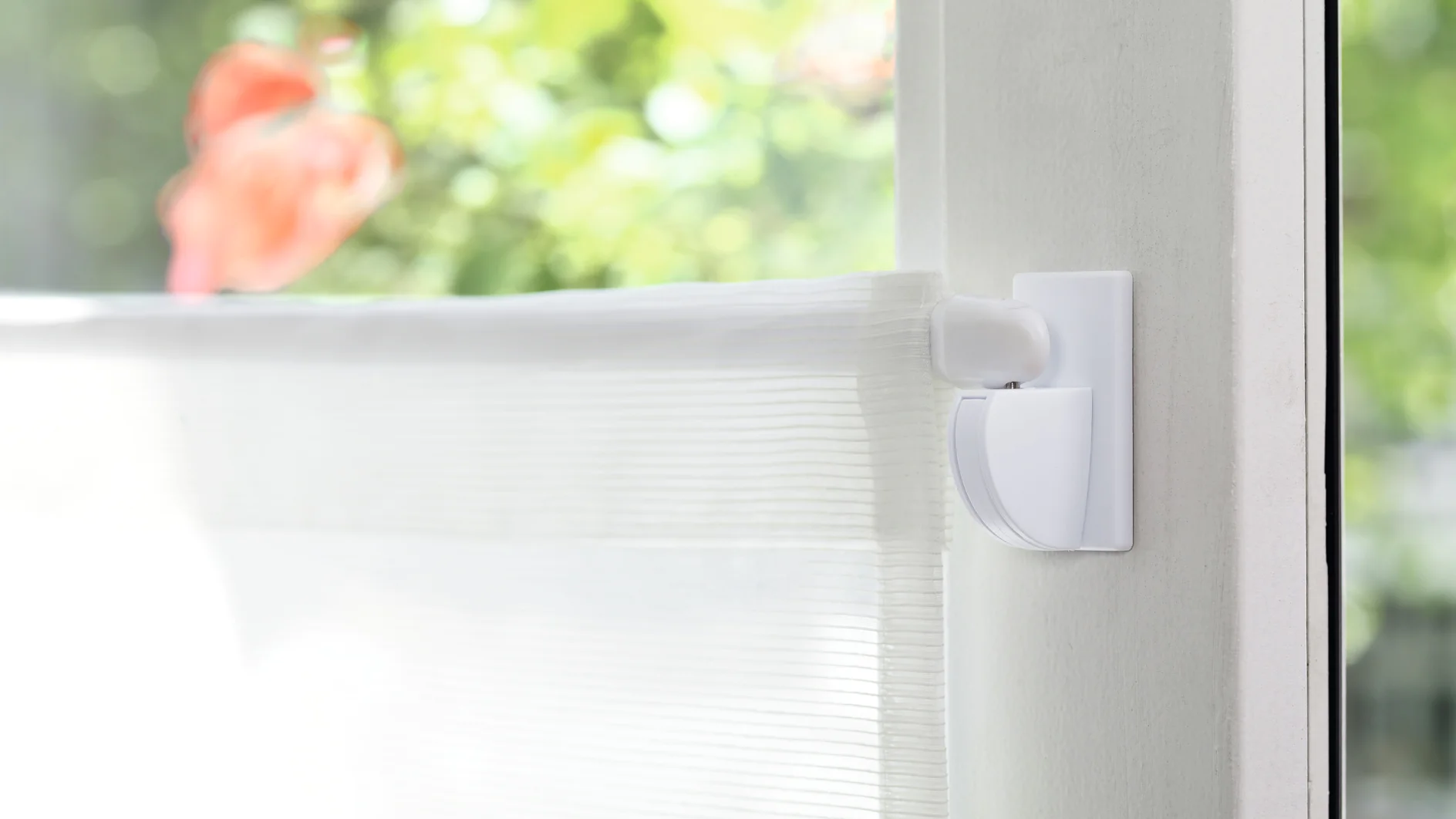 A close-up view of a translucent white window blind partially lowered on a window. The window frame is white, and theres a green blurred garden scene with orange flowers visible outside. Light shines through the blind, creating a soft glow. (This text has been generated by AI)