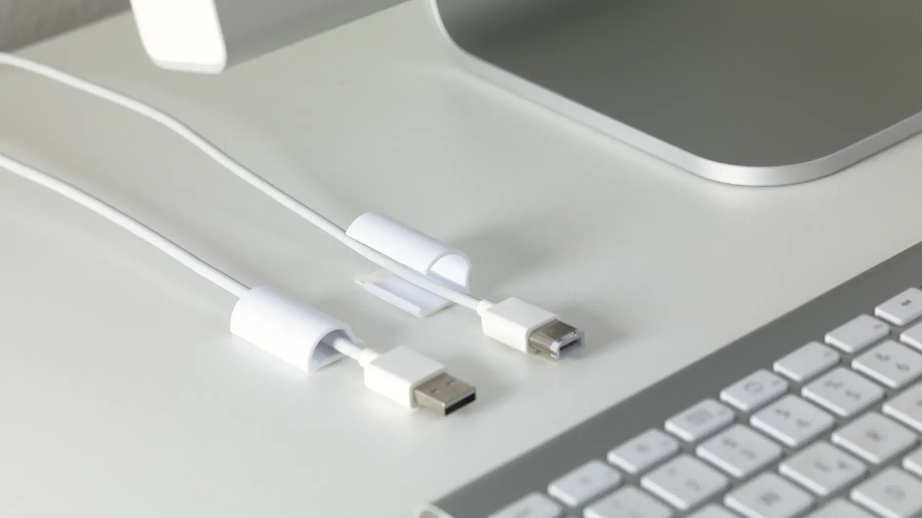 A close-up of a white desk with a corner of a computer monitor and a silver keyboard. Two white USB cables, each held in place by a white clip made with tesa tape, are on the desk next to the keyboard. The scene is minimalistic and tidy. (This text has been generated by AI)