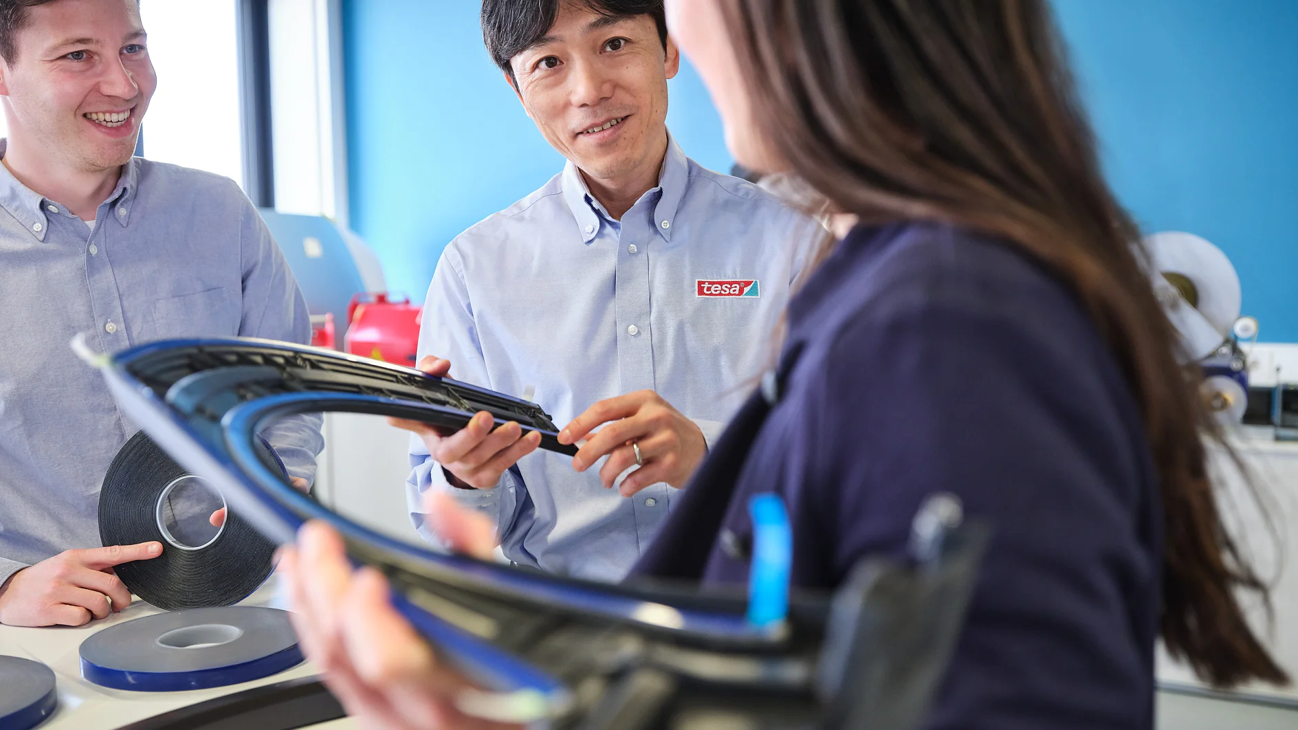 tesa-automotive-key-visual-exterior-005-300dpi Three people in a workspace examine a curved automotive part. Two men in light blue shirts converse with a woman in a dark blazer. Rolls of tesa tape are on the table nearby. The background features a blue wall and a window. (This text has been generated by AI)
