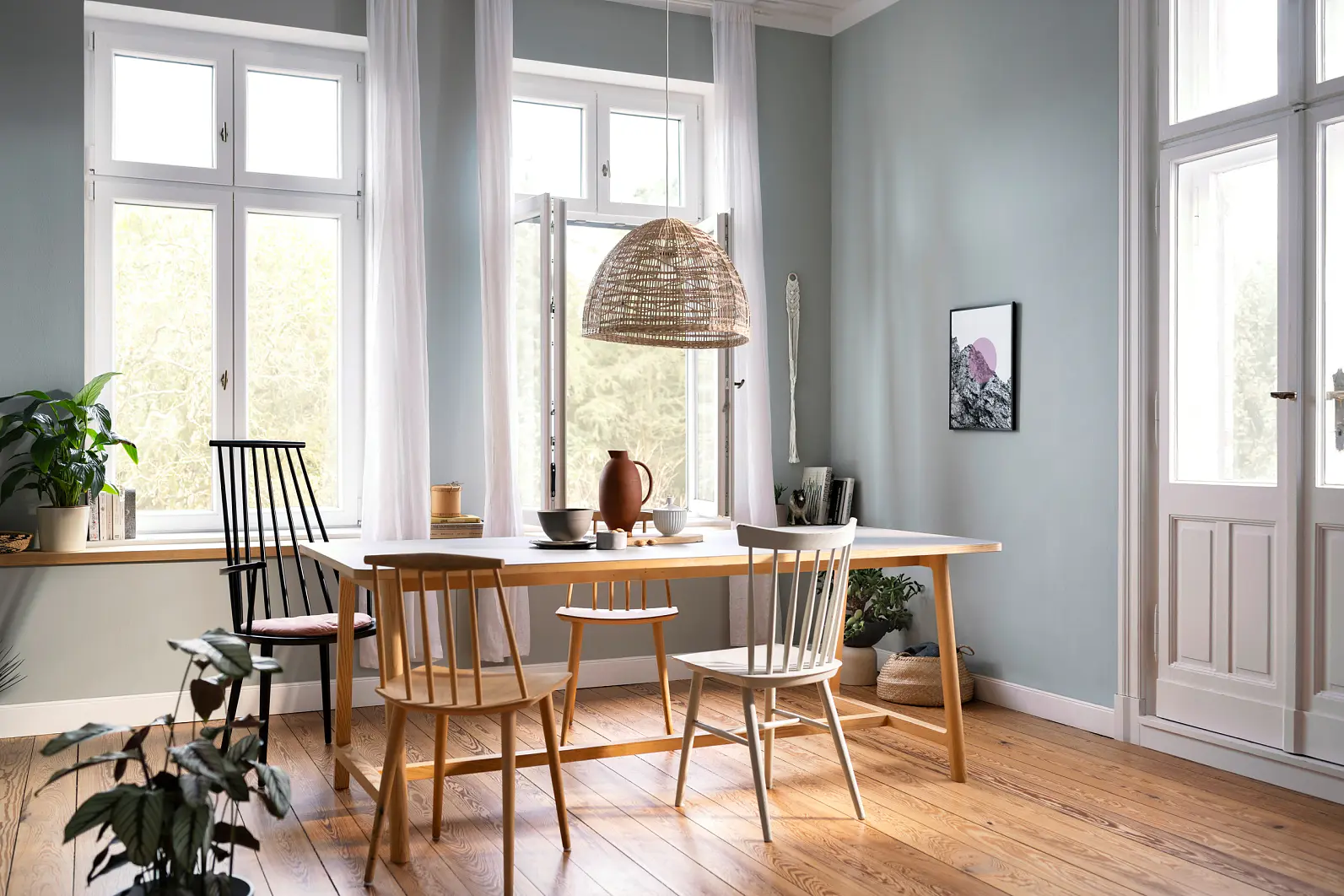 A dining room with a wooden table and six chairs, some mismatched. A woven light fixture hangs above. Large windows let in natural light. The room features light blue walls, wood flooring, and plants in the foreground and on the windowsill. (This text has been generated by AI)
