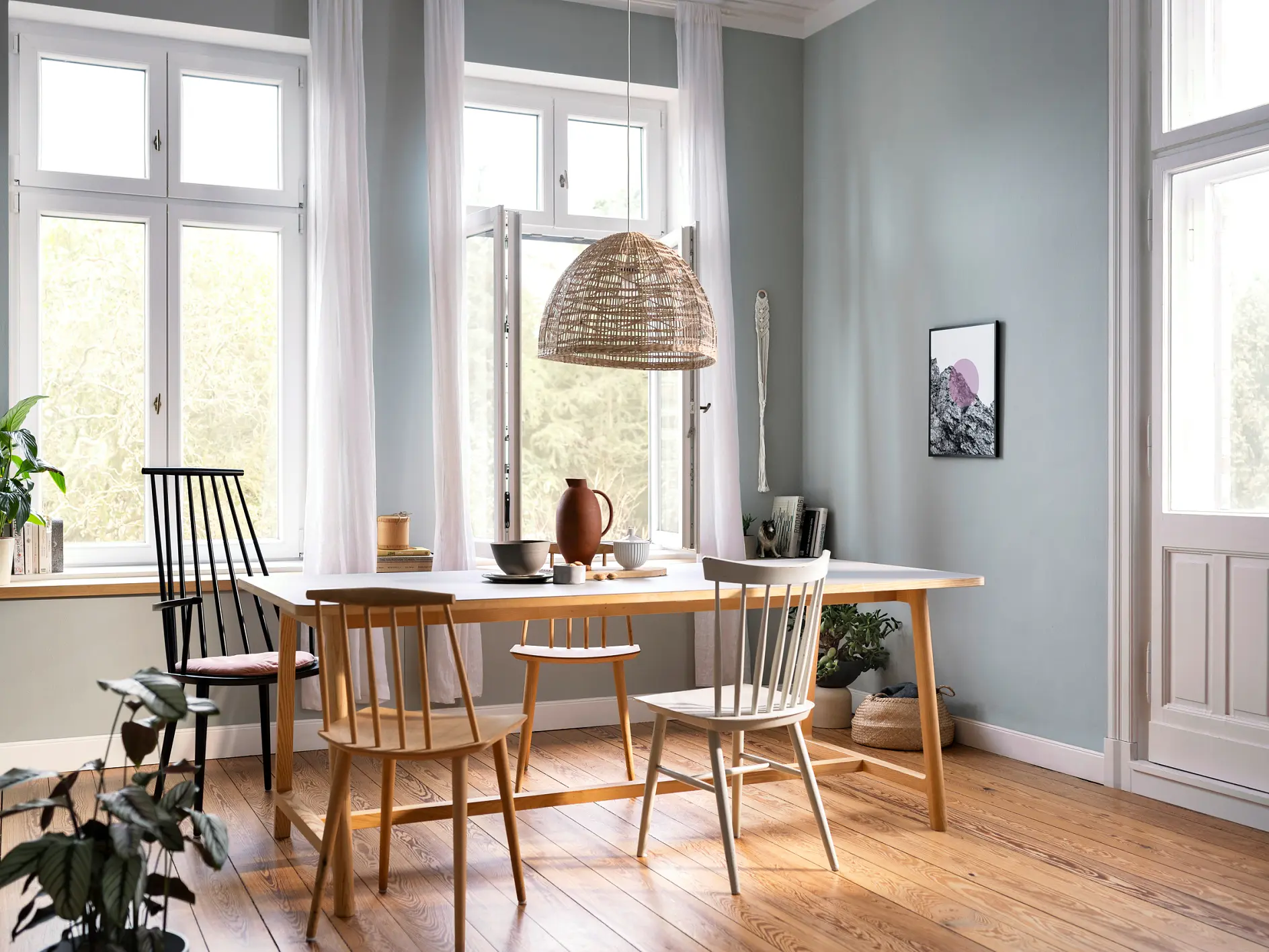 A dining room with a wooden table and six chairs, some mismatched. A woven light fixture hangs above. Large windows let in natural light. The room features light blue walls, wood flooring, and plants in the foreground and on the windowsill.