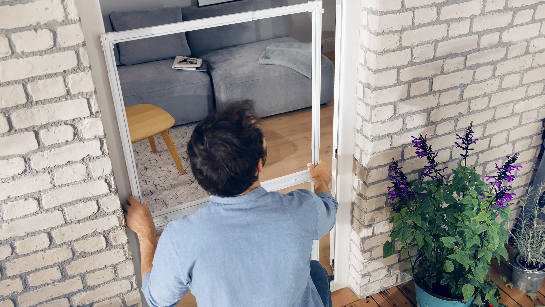 A person in a blue shirt is installing a white doorframe in a room using tesa tape. The floor is wooden, and the walls are brick. To the right, there is a potted plant with purple flowers. In the background, theres a couch and a small wooden table.