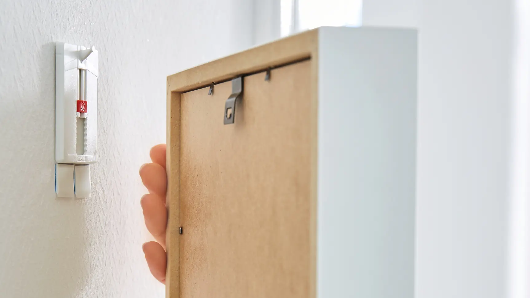 A person is hanging a rectangular picture frame on a white wall using a removable adhesive strip hook made with tesa tape. The wall has a subtle textured pattern, and the frame is being aligned with the hook.