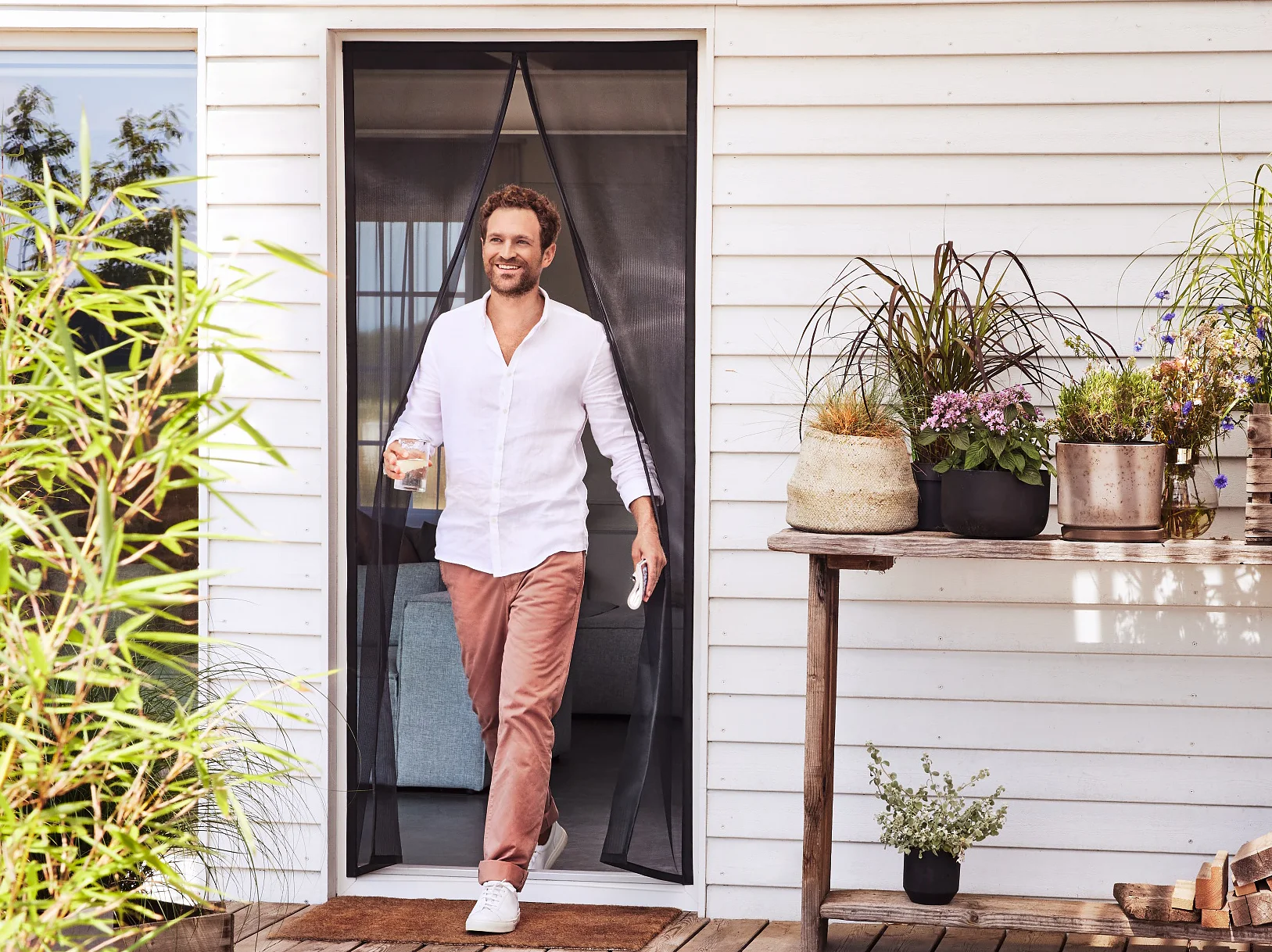 A man wearing a white shirt and pink pants walks through a door onto a wooden deck, holding a glass. The deck features potted plants, a stack of firewood, and a table with more plants. The building has white siding. (This text has been generated by AI)
