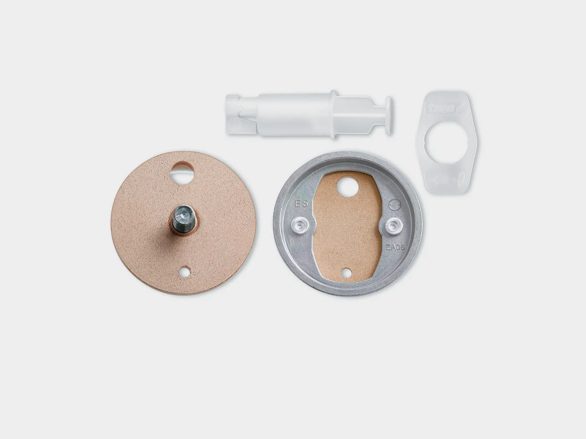 A disassembled rotary dimmer switch kit on a white background. Pieces include a beige round cover, a metal plate with screw holes, a white cylindrical component, and a rectangular plastic piece with a circular cutout. Theres also tesa tape included for secure assembly. (This text has been generated by AI)
