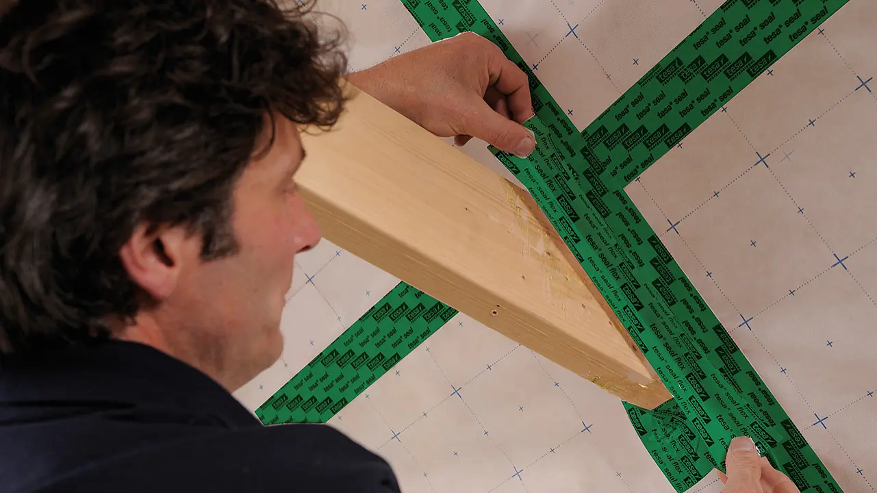 A person applies green tesa tape over overlapping sections of material, sealing them. They hold a wooden beam in one hand, positioned across the taped area. The background shows a grid pattern on the material. (This text has been generated by AI)