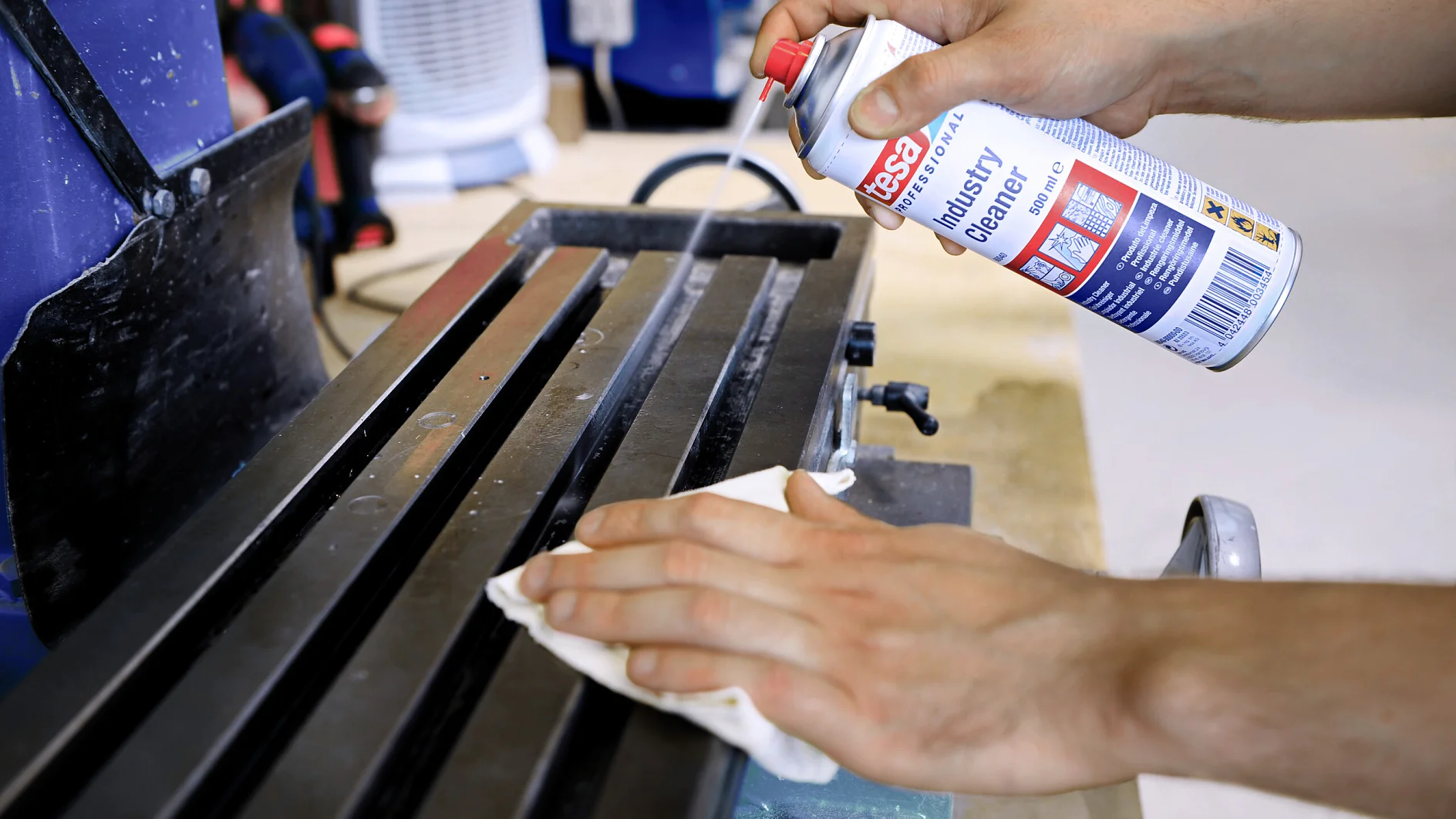 Eine Person reinigt mit einem weißen Tuch und einer Aerosoldose mit der Aufschrift „Industriereiniger“ die Metalloberfläche einer Maschine. Im Hintergrund sind eine blaue Maschine und ein weißer Heizer zu sehen. (This text has been generated by AI)