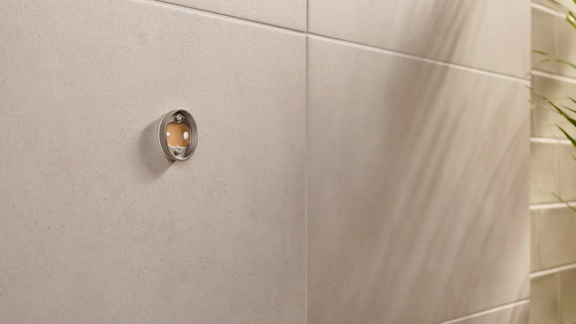 Close-up of a beige tiled wall with a circular metal fitting. The fitting has a transparent front, revealing a small reddish-brown circle inside. On the right, the shadow of a plant is visible on the wall.