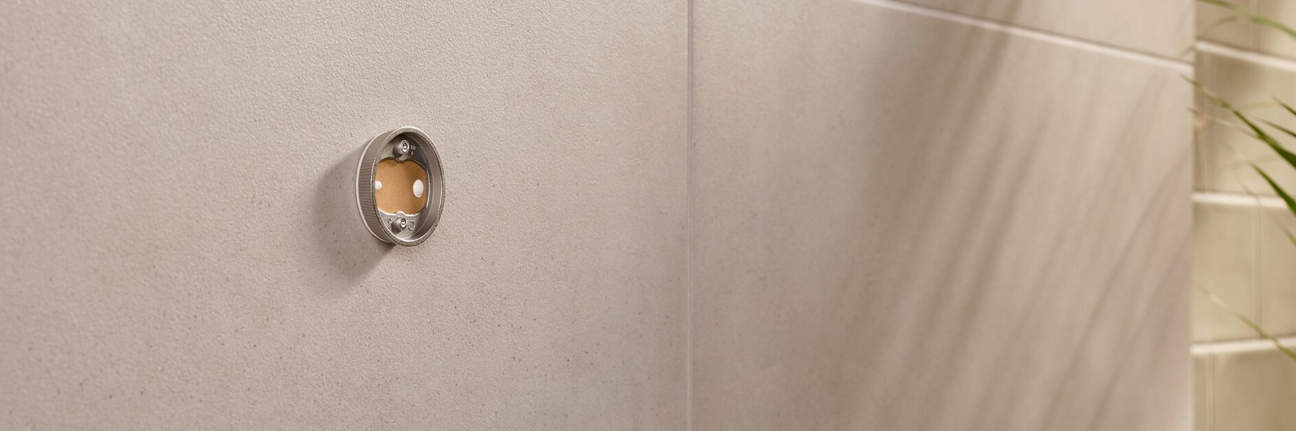 Close-up of a beige tiled wall with a circular metal fitting. The fitting has a transparent front, revealing a small reddish-brown circle inside. On the right, the shadow of a plant is visible on the wall.