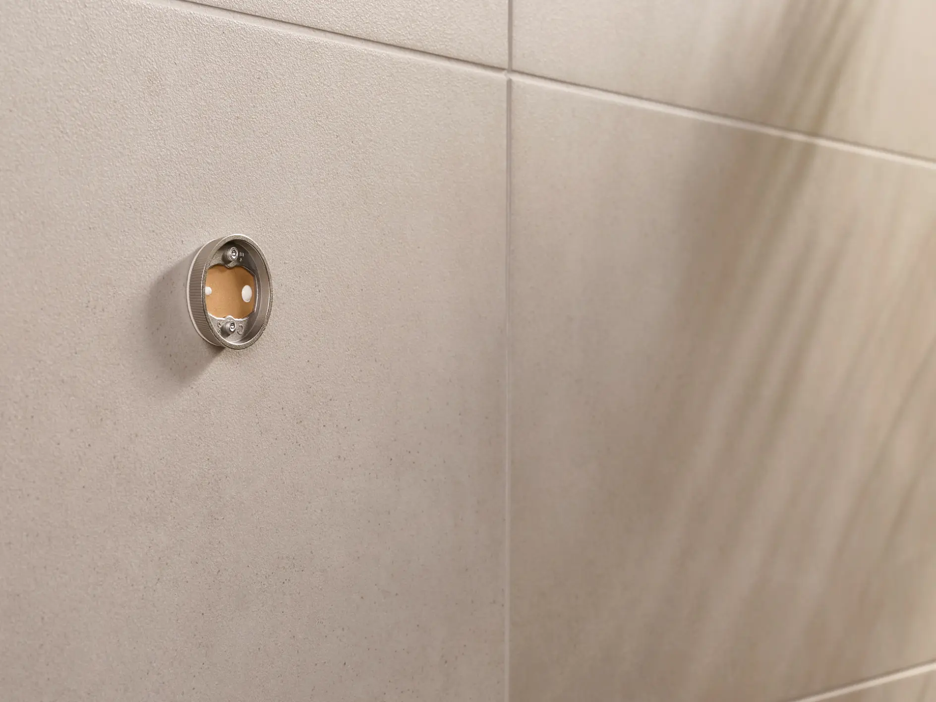 Close-up of a beige tiled wall with a circular metal fitting. The fitting has a transparent front, revealing a small reddish-brown circle inside. On the right, the shadow of a plant is visible on the wall.