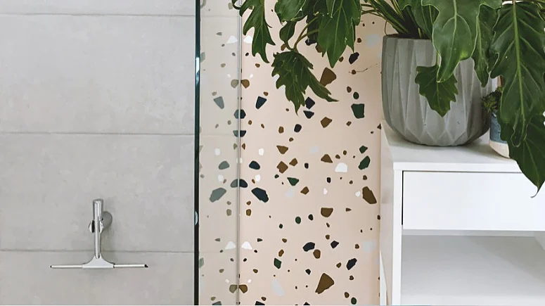 kleben-statt-bohren-badOkkXKUqwQN8ah_800x800 Ein Badezimmer verfügt über eine moderne Terrazzo-Duschwand mit bunten Fragmenten. Daneben steht auf einem weißen Schrank ein Topf mit üppigem grünem Blattwerk. Das Gesamtdesign ist minimalistisch und frisch. (This text has been generated by AI)
