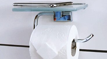 An einer Wand ist ein verchromter Toilettenpapierhalter angebracht, auf dem sich eine Rolle weißes Tesa-Klebeband befindet. Darüber liegt auf einer kleinen Glasablage ein Smartphone. Die Wand ist weiß mit schwarzen Fugen. (This text has been generated by AI)