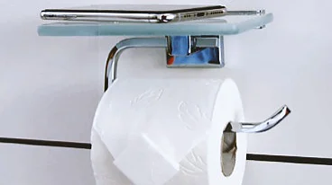 An einer Wand ist ein verchromter Toilettenpapierhalter angebracht, auf dem sich eine Rolle weißes Tesa-Klebeband befindet. Darüber liegt auf einer kleinen Glasablage ein Smartphone. Die Wand ist weiß mit schwarzen Fugen. (This text has been generated by AI)