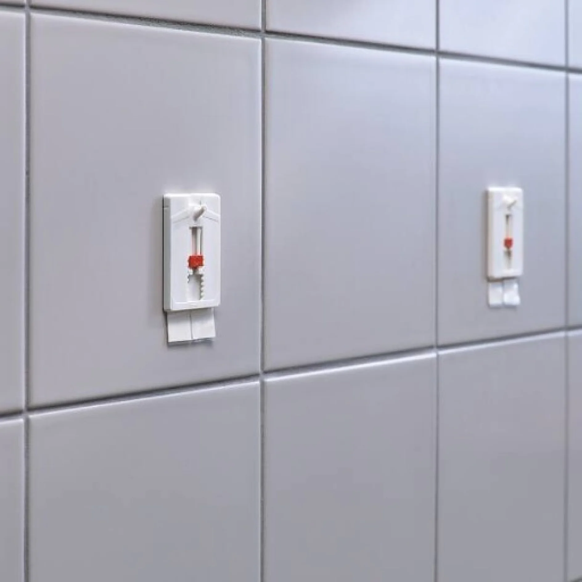 Grau geflieste Wand mit drei roten Fliesenabstandshaltern, die mit Tesa-Klebeband befestigt sind. Die Abstandshalter sorgen für einen gleichmäßigen Abstand und eine gleichmäßige Ausrichtung der Fliesen. Die Fliesen haben eine reflektierende Oberfläche und subtile Farbvariationen. (This text has been generated by AI)