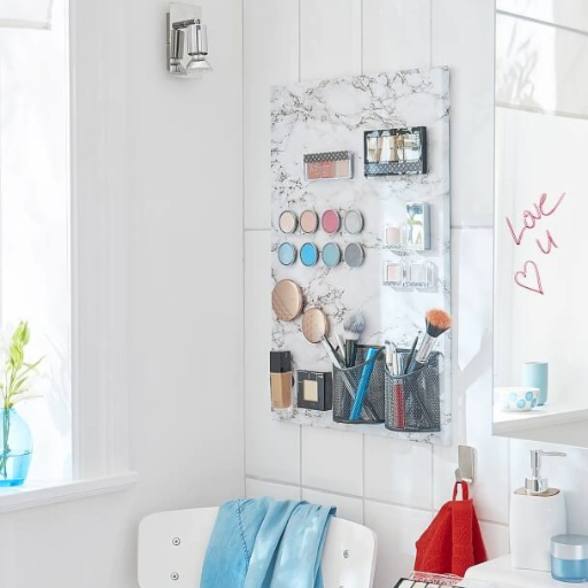 In einer Badezimmerszene mit einer weiß gefliesten Wand ist ein Make-up-Organizer mit Kosmetika und Pinseln zu sehen. Ein Handtuch hängt an einem Haken und auf dem Spiegel steht mit Tesa-Klebeband in rotem Lippenstift „Love U“. Auf der Fensterbank ist eine kleine Pflanze zu sehen. (This text has been generated by AI)