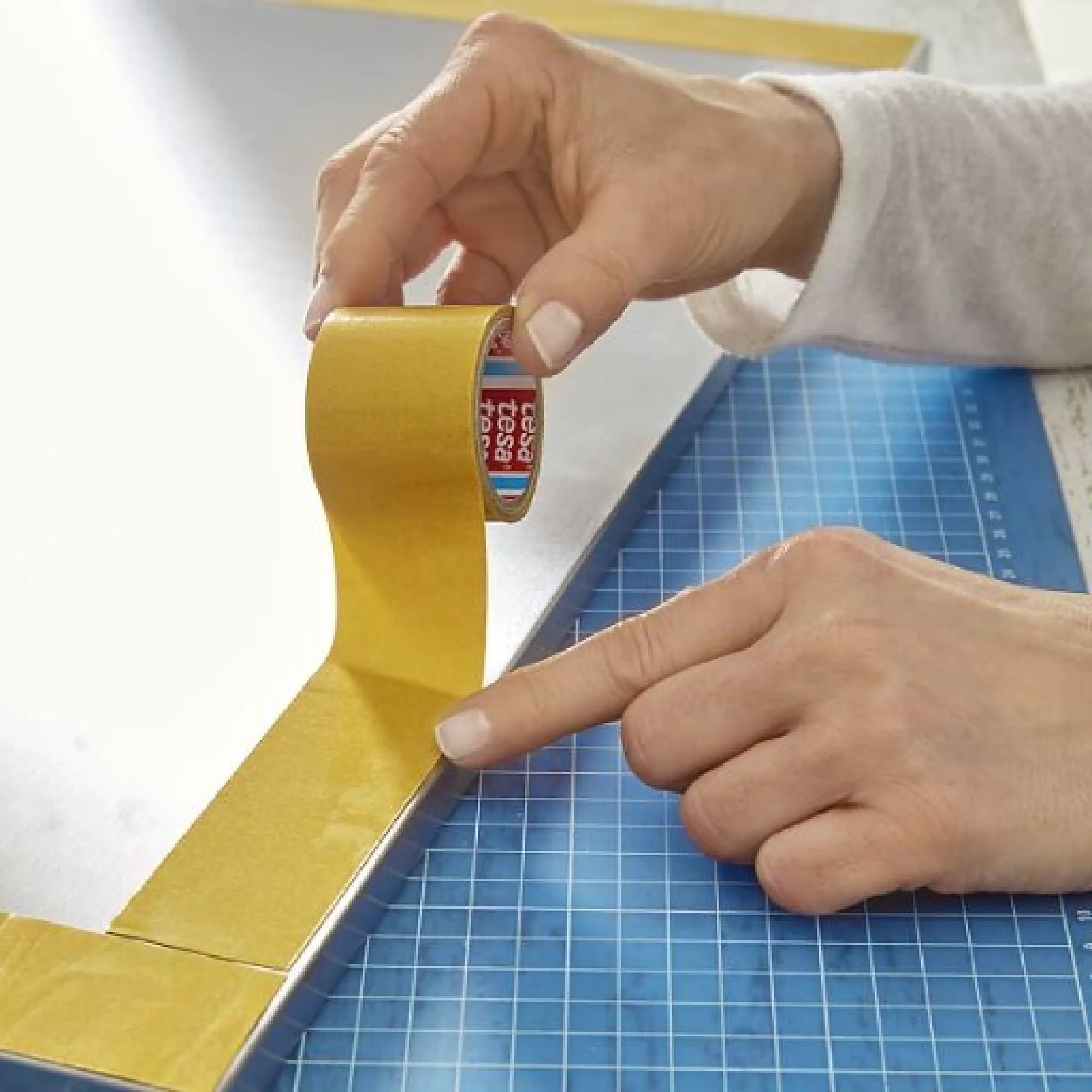 sms_makeUpBoard_007_1x1 Eine Person klebt goldenes Tesaband auf die Kante einer großen, flachen Oberfläche. Das Tesaband wird von einer Rolle abgewickelt, die sie in einer Hand hält. Unter der Oberfläche ist eine blaue Schneidematte mit einem Gittermuster zu sehen. (This text has been generated by AI)