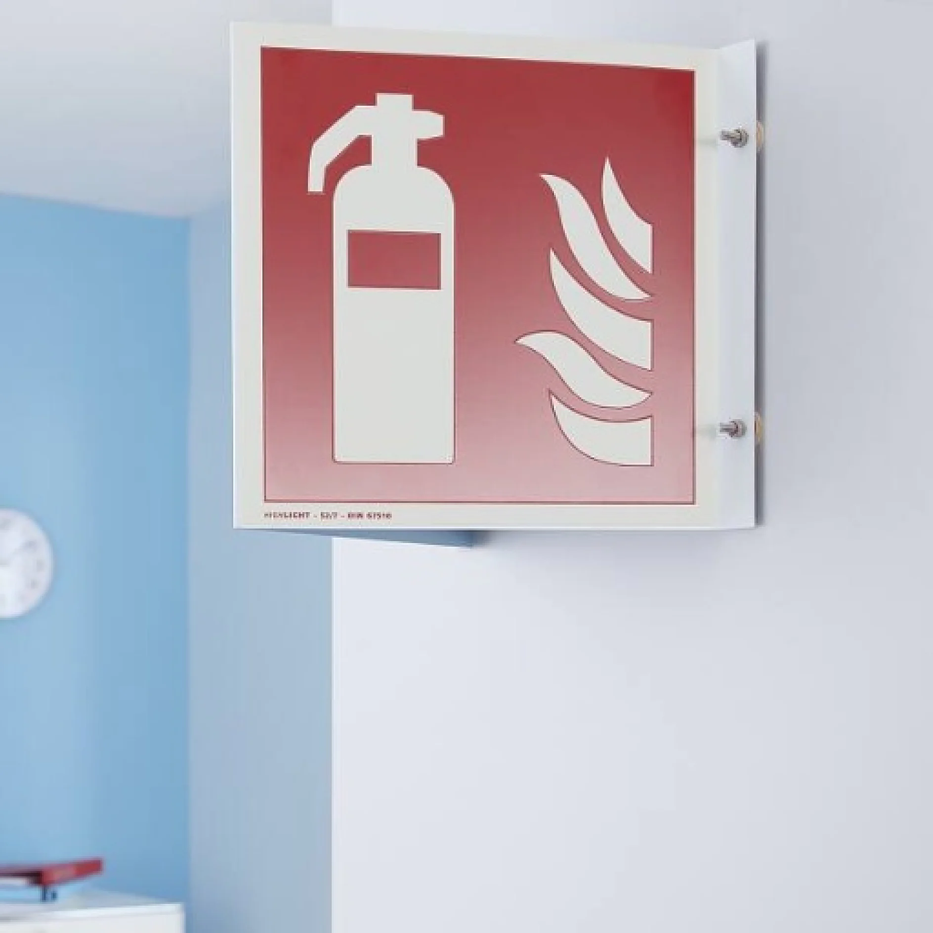 Ein rotes Feuerlöscherschild mit weißer Grafik ist mit Tesa-Klebeband an einer Wand befestigt. Es zeigt ein Feuerlöscher-Symbol und Flammensymbole. Die Wand ist hellblau und im Hintergrund ist eine Uhr zu sehen. (This text has been generated by AI)