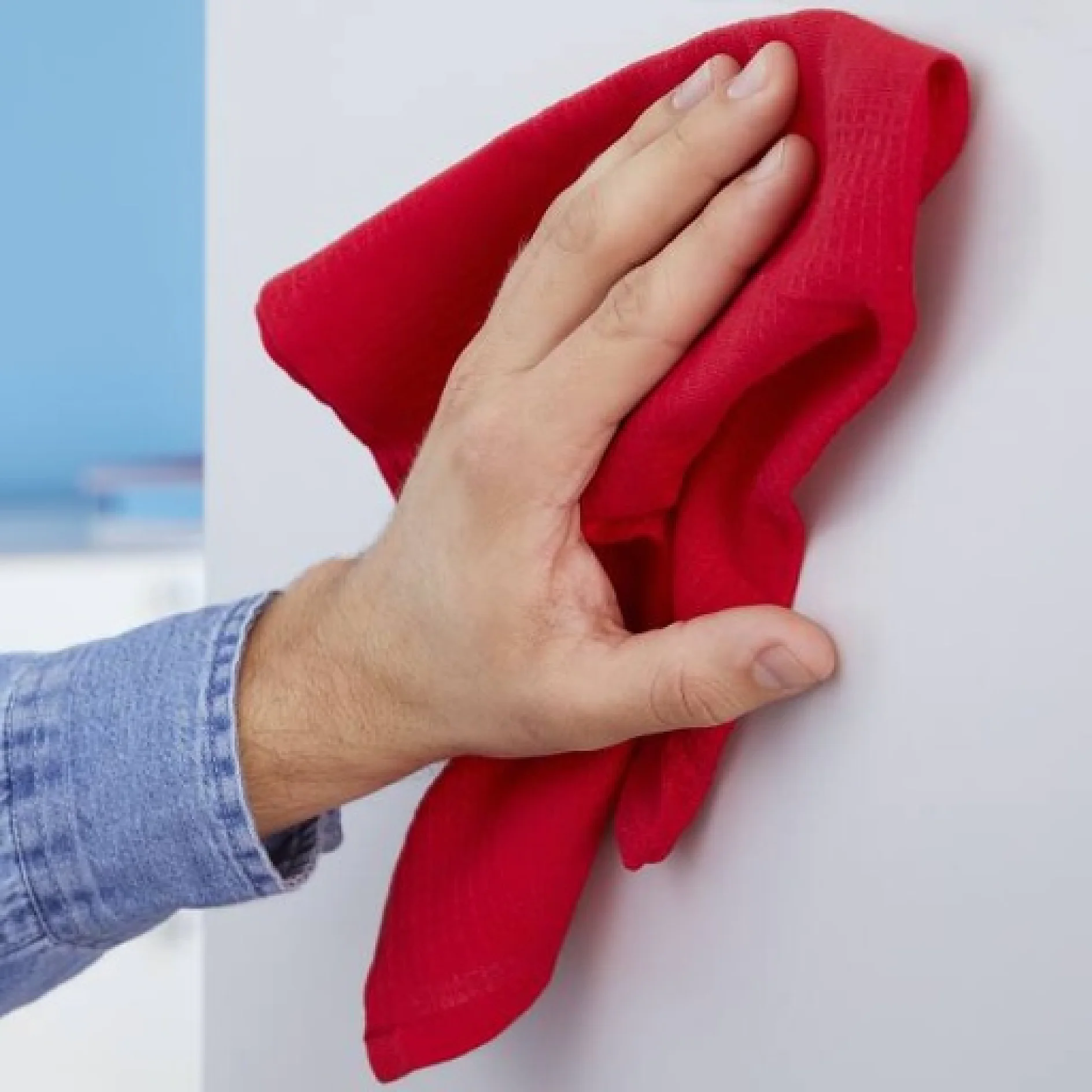 tesa_77901_adhesive_screw_sign_step_1of8_ap_1x1 Eine Hand mit einem blauen Ärmel mit Manschette wischt mit einem roten Tuch eine weiße Oberfläche ab. Der Hintergrund ist verschwommen und zeigt eine hellblaue Wand und die Andeutung eines Regals. (This text has been generated by AI)