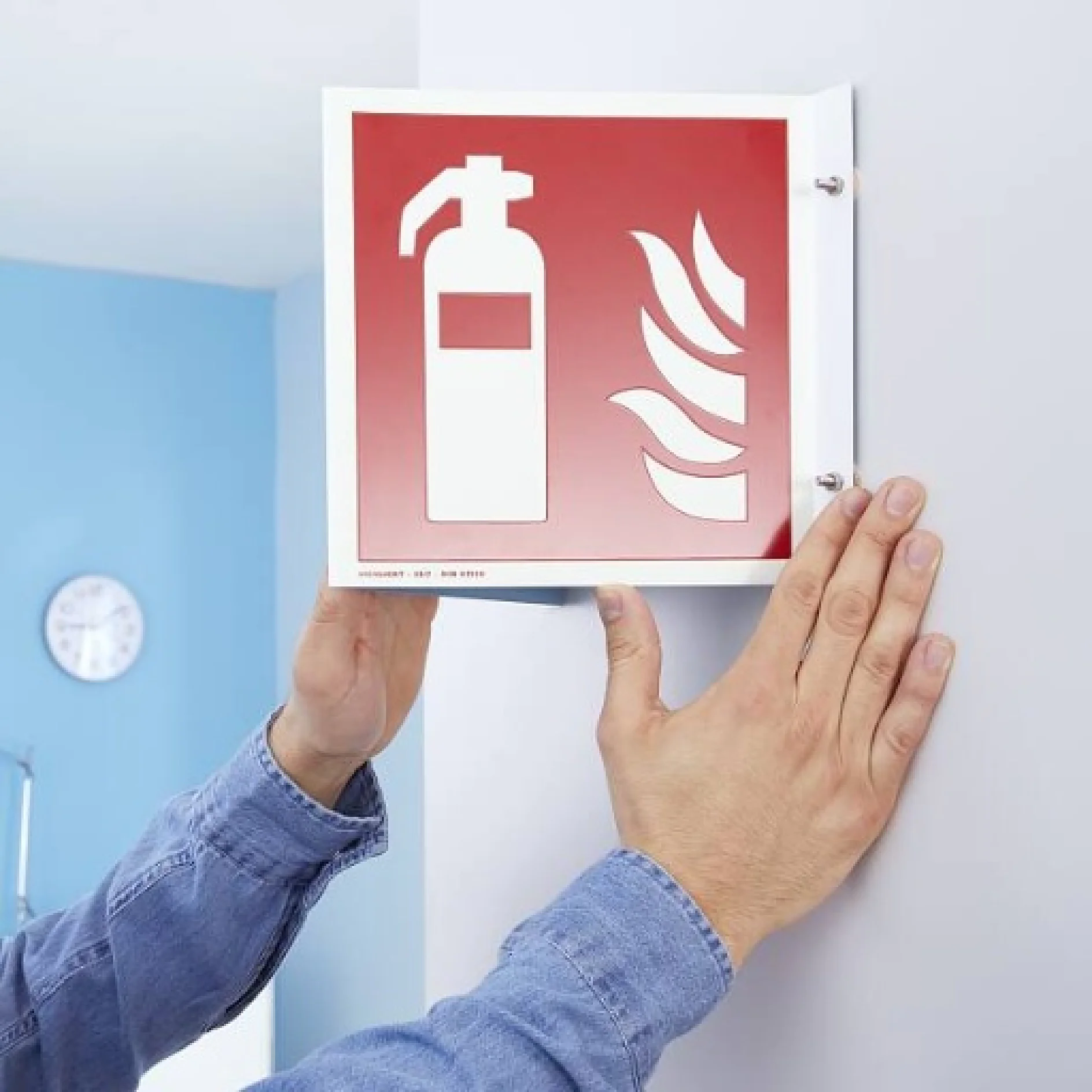 Eine Person befestigt ein rotes Feuerlöscher-Schild an einer weißen Wand. Das Schild zeigt ein weißes Feuerlöscher-Symbol und Flammen. Die Person befestigt es mit beiden Händen und verwendet dabei Tesa-Klebeband. Im Hintergrund ist eine Uhr an einer hellblauen Wand zu sehen. (This text has been generated by AI)