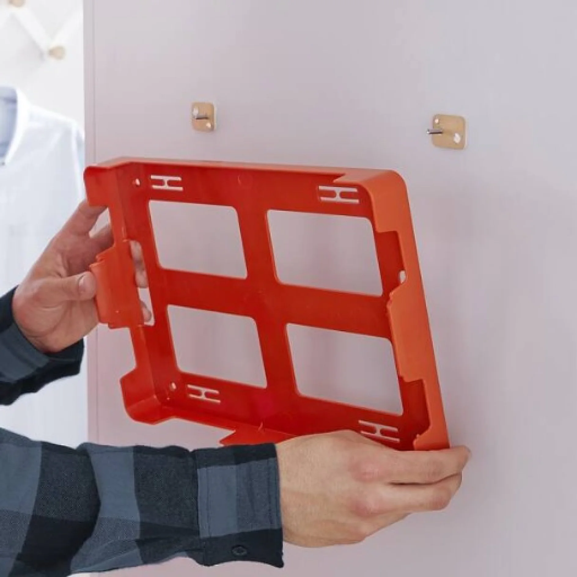 Eine Person im karierten Hemd hängt einen orangefarbenen Organizer aus Kunststoff mit Tesa-Klebeband an eine weiße Wand. An der Wand sind zwei Metallhaken zu sehen, die den Organizer halten. (This text has been generated by AI)