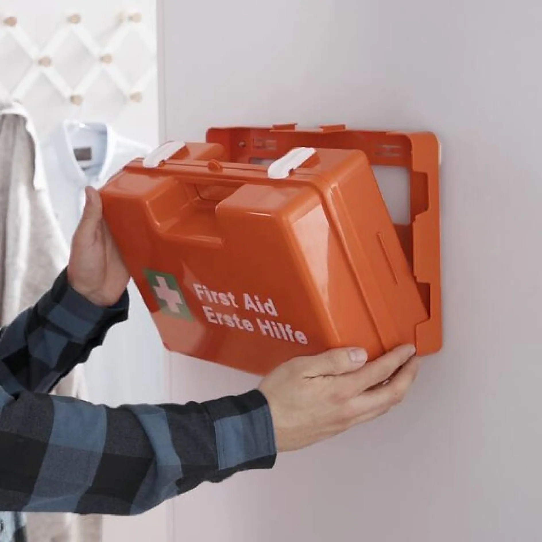 Eine Person im karierten Hemd befestigt einen orangefarbenen Verbandskasten mit der Aufschrift „First Aid Erste Hilfe“ mit Tesa-Klebeband an einer Wandhalterung. Im Hintergrund ist unscharf die Schulter einer weiteren Person zu sehen, darüber hängt ein weißer Kittel. (This text has been generated by AI)
