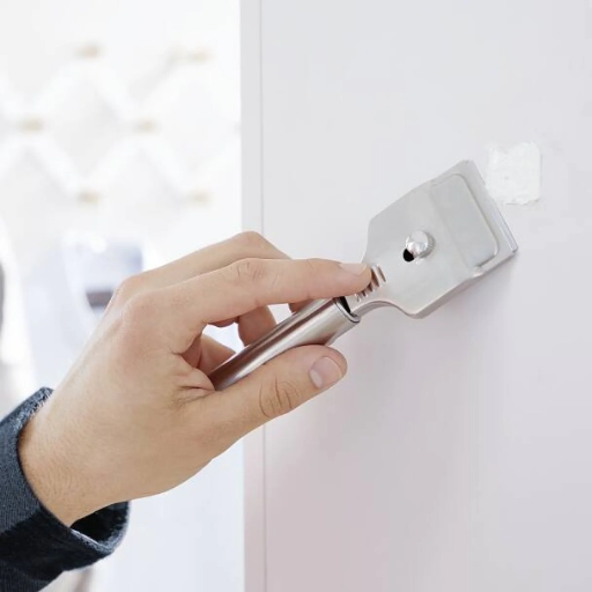 Eine Hand glättet mit einem Metallschaber die Oberfläche einer weißen Wand. Auf der Wand befindet sich ein kleiner Fleck weißer Kitt oder Spachtelmasse, der mit Tesa-Klebeband befestigt ist. Der Hintergrund ist unscharf und konzentriert sich auf die Hand und das Werkzeug. (This text has been generated by AI)