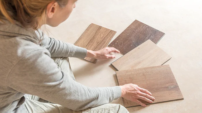 Eine Person in einem grauen Kapuzenpullover sitzt auf dem Boden, betrachtet vier Holzbodenmuster in verschiedenen Farbtönen und Texturen und ordnet sie. Die Oberfläche darunter ist ein schlichter, heller Boden. Sie verwenden Tesa-Klebeband, um jedes Muster sorgfältig an Ort und Stelle zu befestigen, während sie die Optionen vergleichen. (This text has been generated by AI)