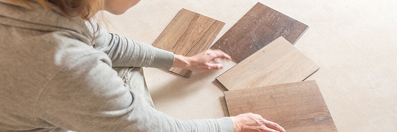 Eine Person in einem grauen Kapuzenpullover sitzt auf dem Boden, betrachtet vier Holzbodenmuster in verschiedenen Farbtönen und Texturen und ordnet sie. Die Oberfläche darunter ist ein schlichter, heller Boden. Sie verwenden Tesa-Klebeband, um jedes Muster sorgfältig an Ort und Stelle zu befestigen, während sie die Optionen vergleichen. (This text has been generated by AI)