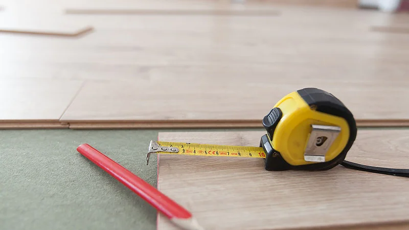 Ein messendes Tesaband und ein roter Bleistift werden auf hellen Holzdielen platziert. Das Tesaband wird über eine der Dielen gezogen und zeigt damit an, dass gerade gemessen wird. Im Hintergrund sind weitere Dielen auf einer hellgrünen Oberfläche zu sehen. (This text has been generated by AI)