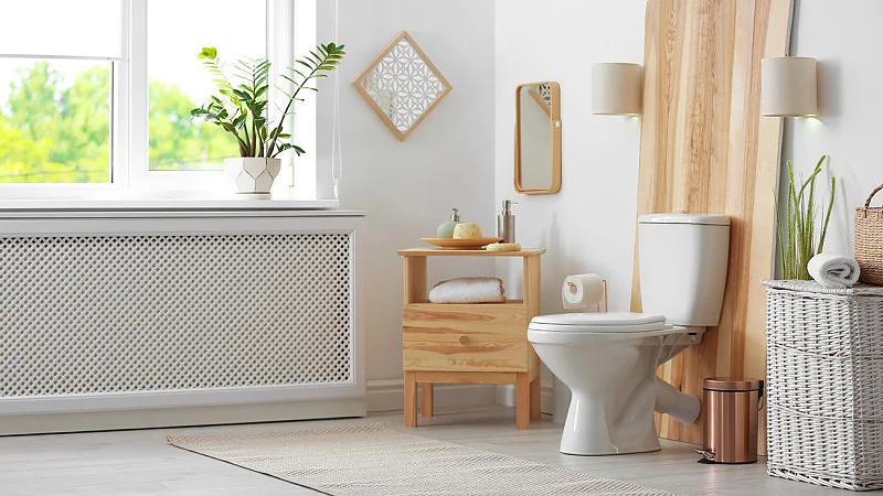 Ein Badezimmer mit einer weißen Toilette vor einer Holzverkleidung, einem kleinen Holzschrank mit Toilettenartikeln, einem weißen Heizkörper und Topfpflanzen. In der Nähe stehen ein Weidenkorb und ein kleiner Mülleimer. Natürliches Licht fällt durch ein Fenster ein. (This text has been generated by AI)